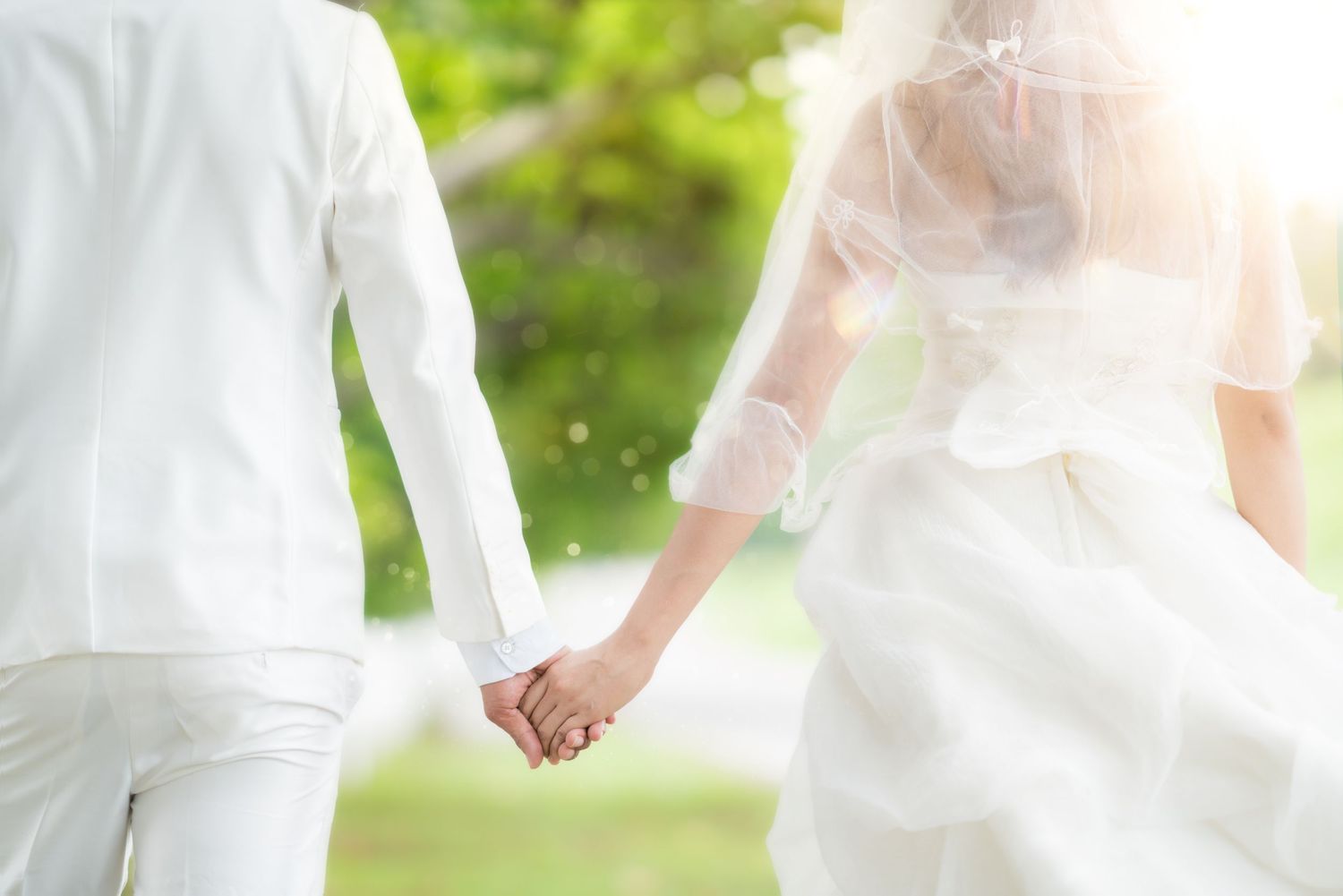 Newlywed couple holding hands in white wedding attire walking through lush green garden.