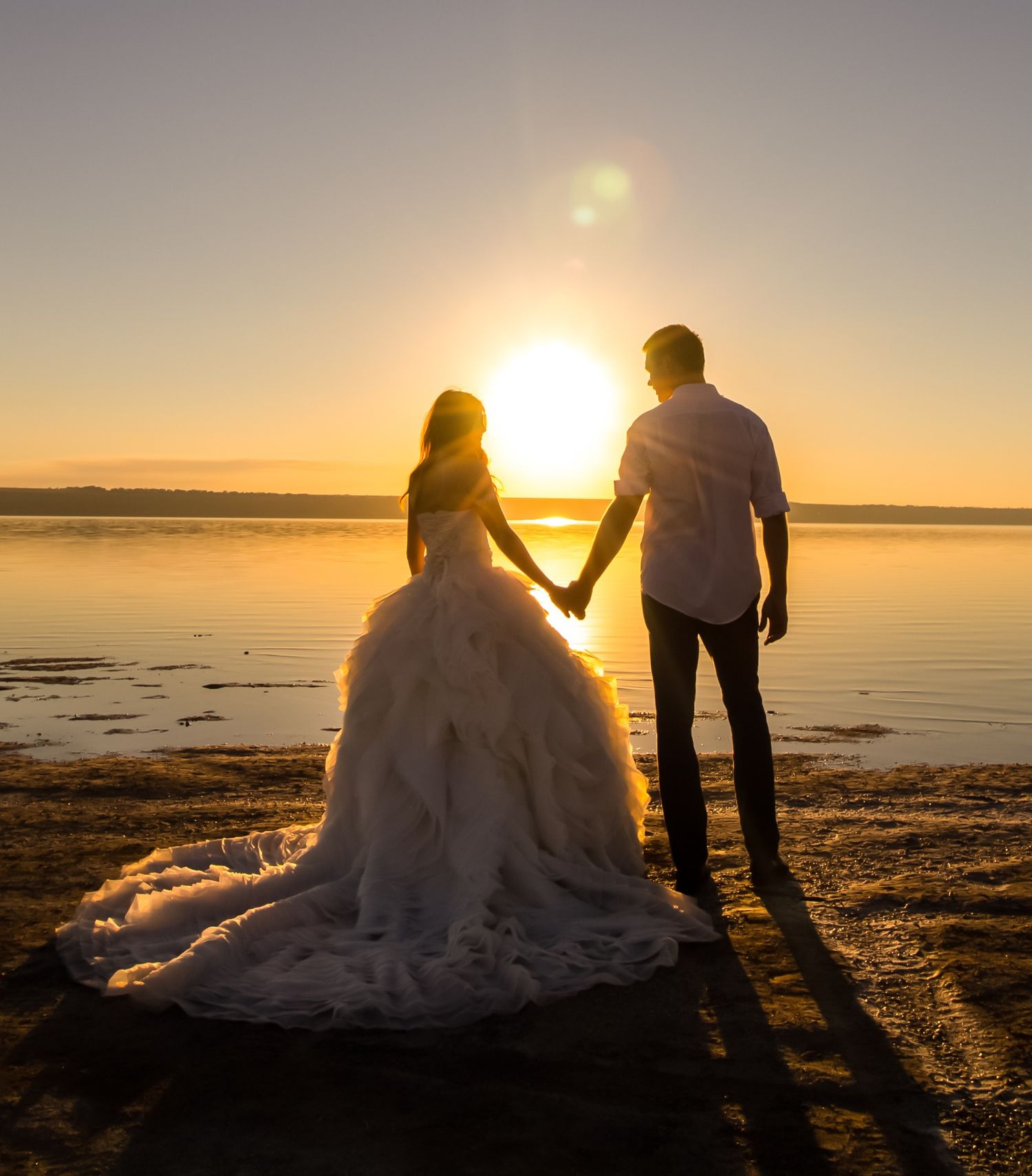 Bride and groom holding hands at sunset on a beach, silhouetted against golden sky in wedding attire.