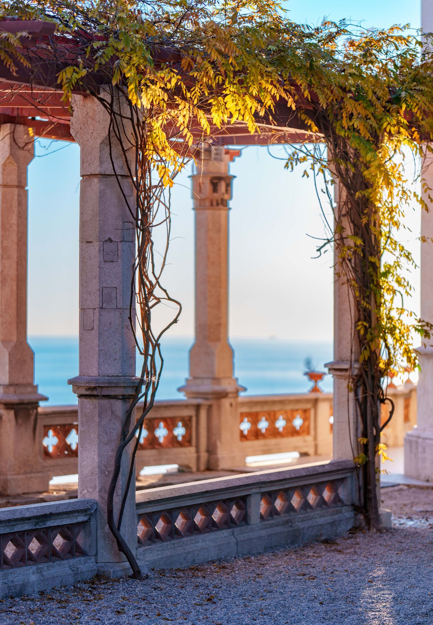 Stone pergola with climbing vines overlooking a serene sea at golden hour, featuring ornate columns and a decorative balustrade.