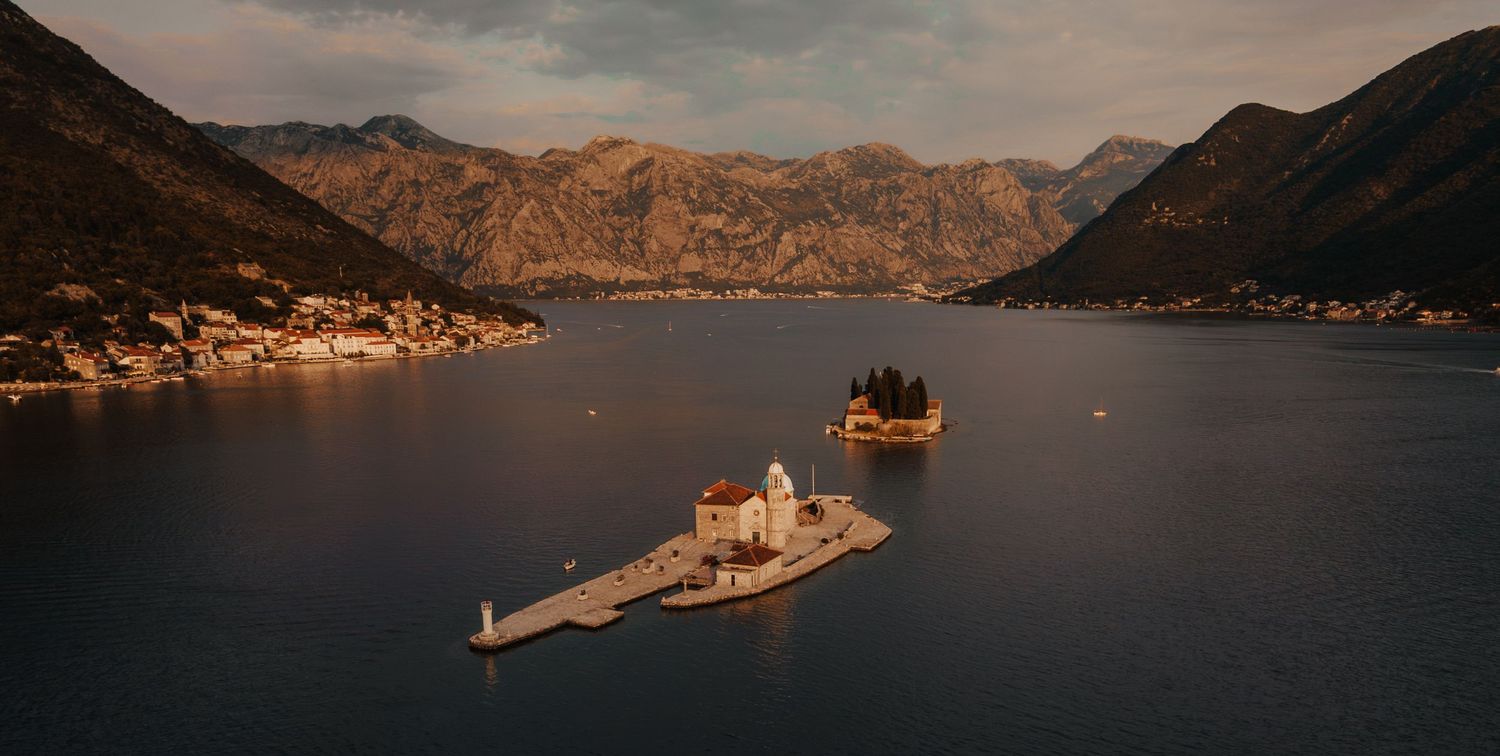 Aerial panoramic view of two small islands with a church on a calm blue bay surrounded by mountains.