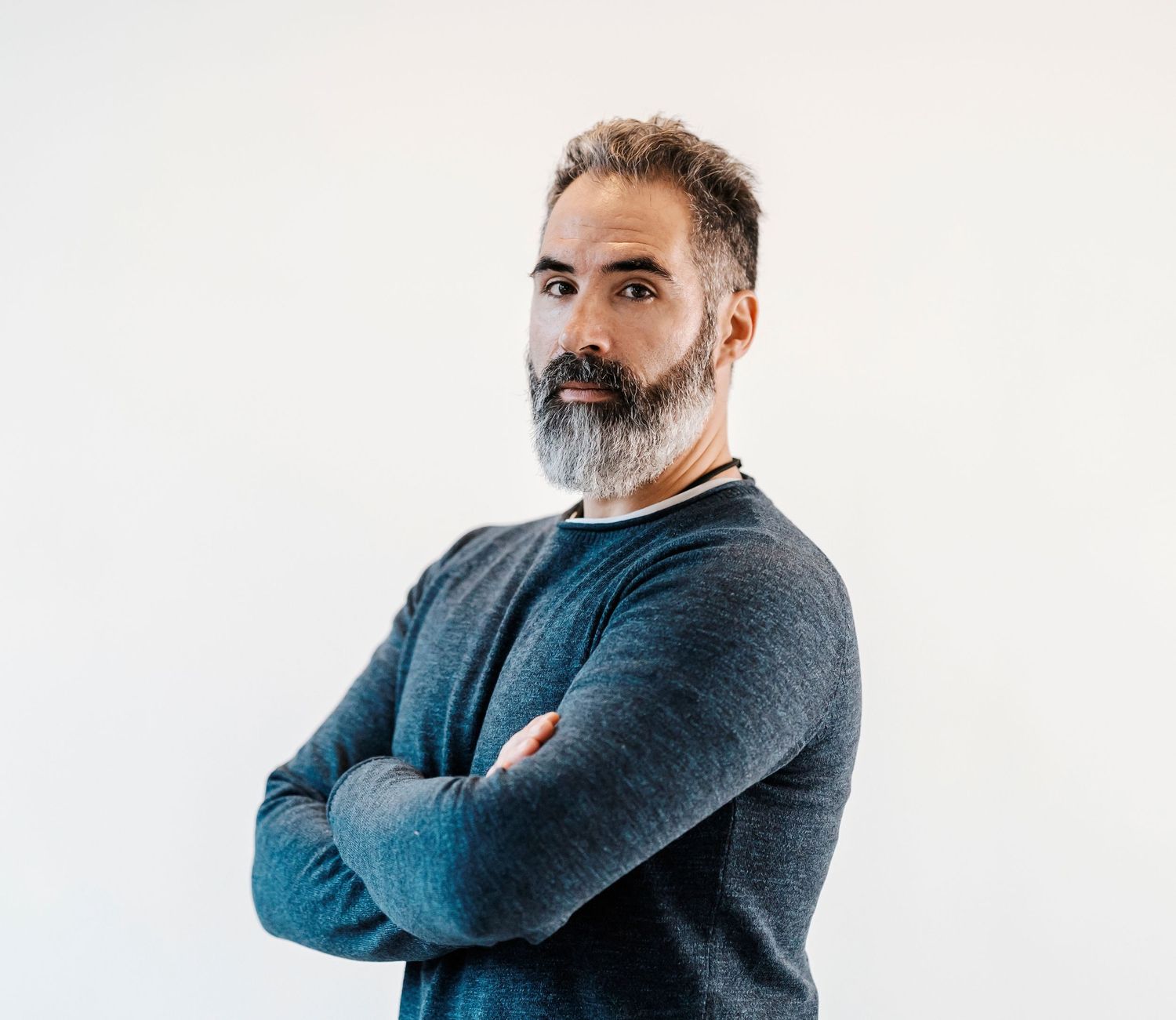 Bearded man with gray hair standing with arms crossed in a blue sweater on white background.
