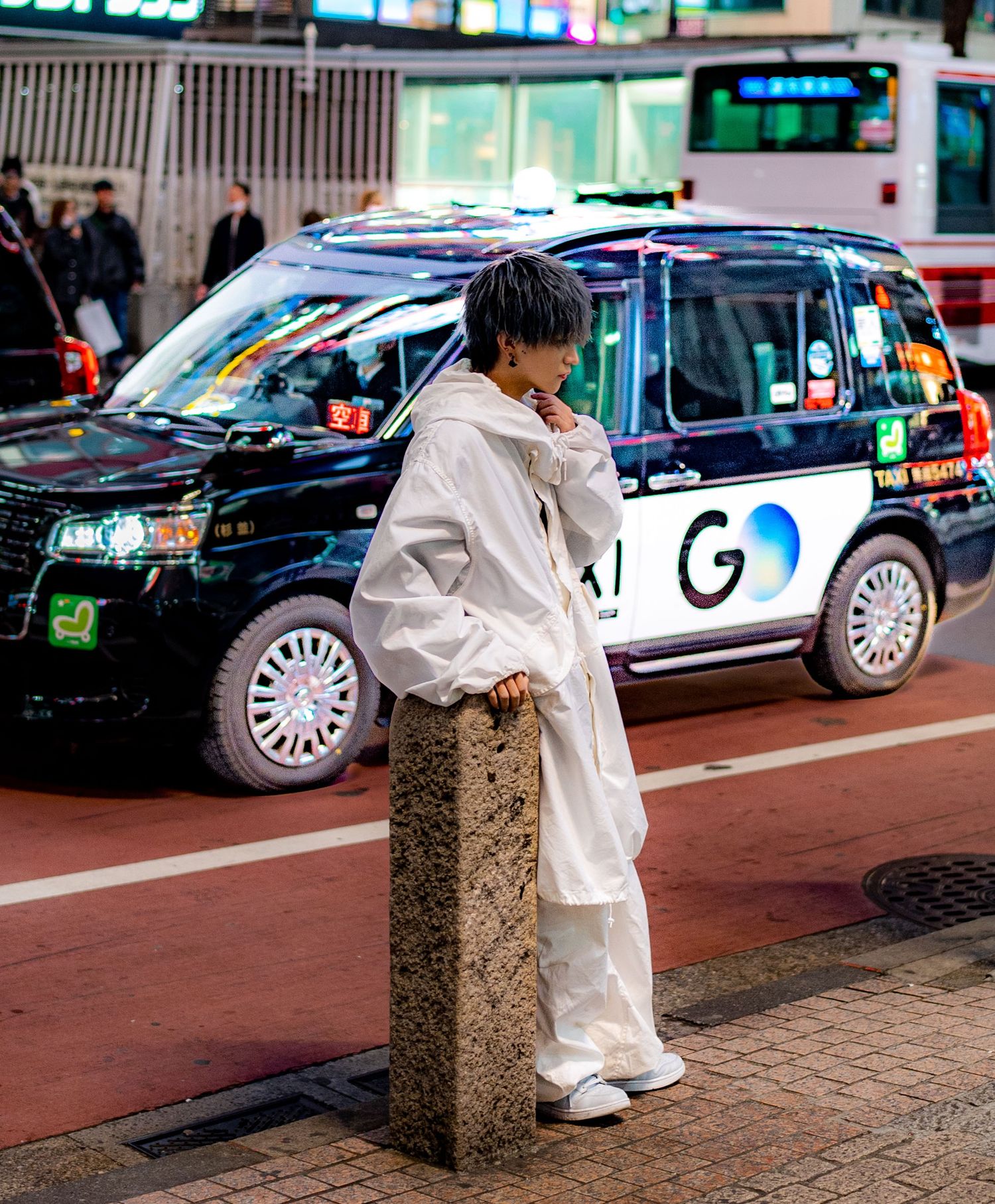 A young person in a white outfit leans on a stone post near a black GO taxi on a busy Japanese street.