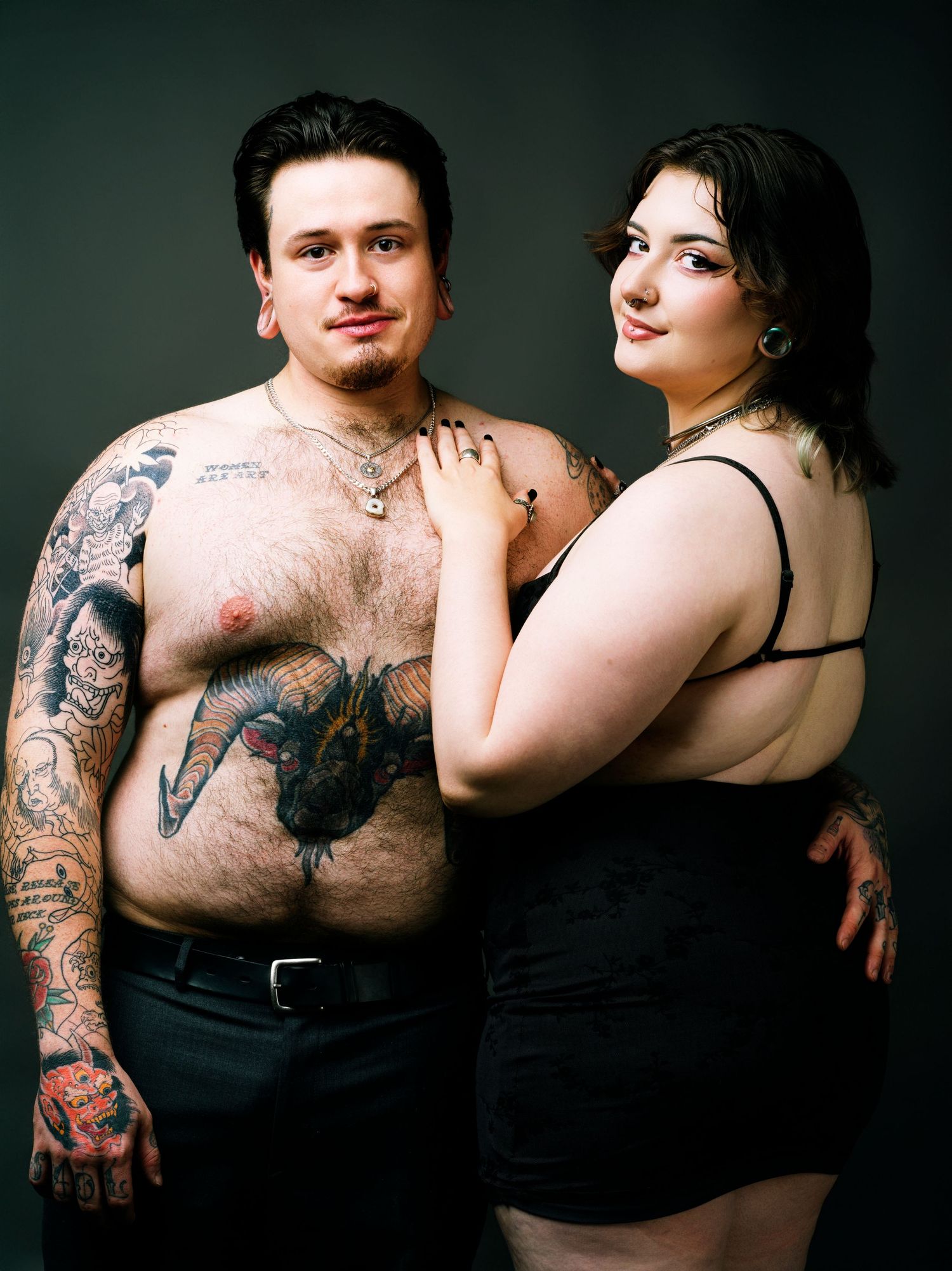 A tattooed shirtless man and a plus-size woman in black pose together for a couples portrait photo.