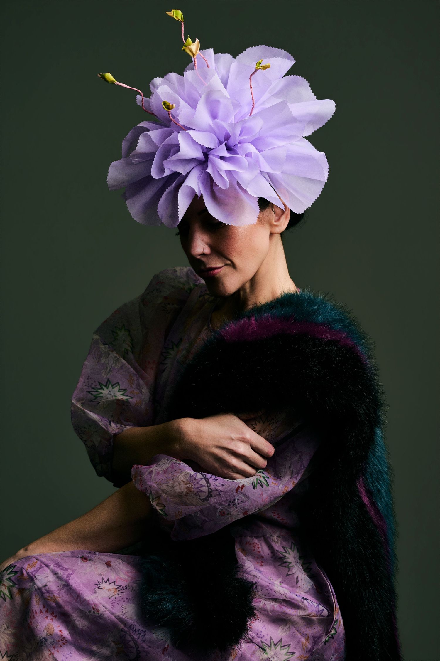 Woman in lavender floral dress wearing large purple flower fascinator hat with fur stole on dark green background.