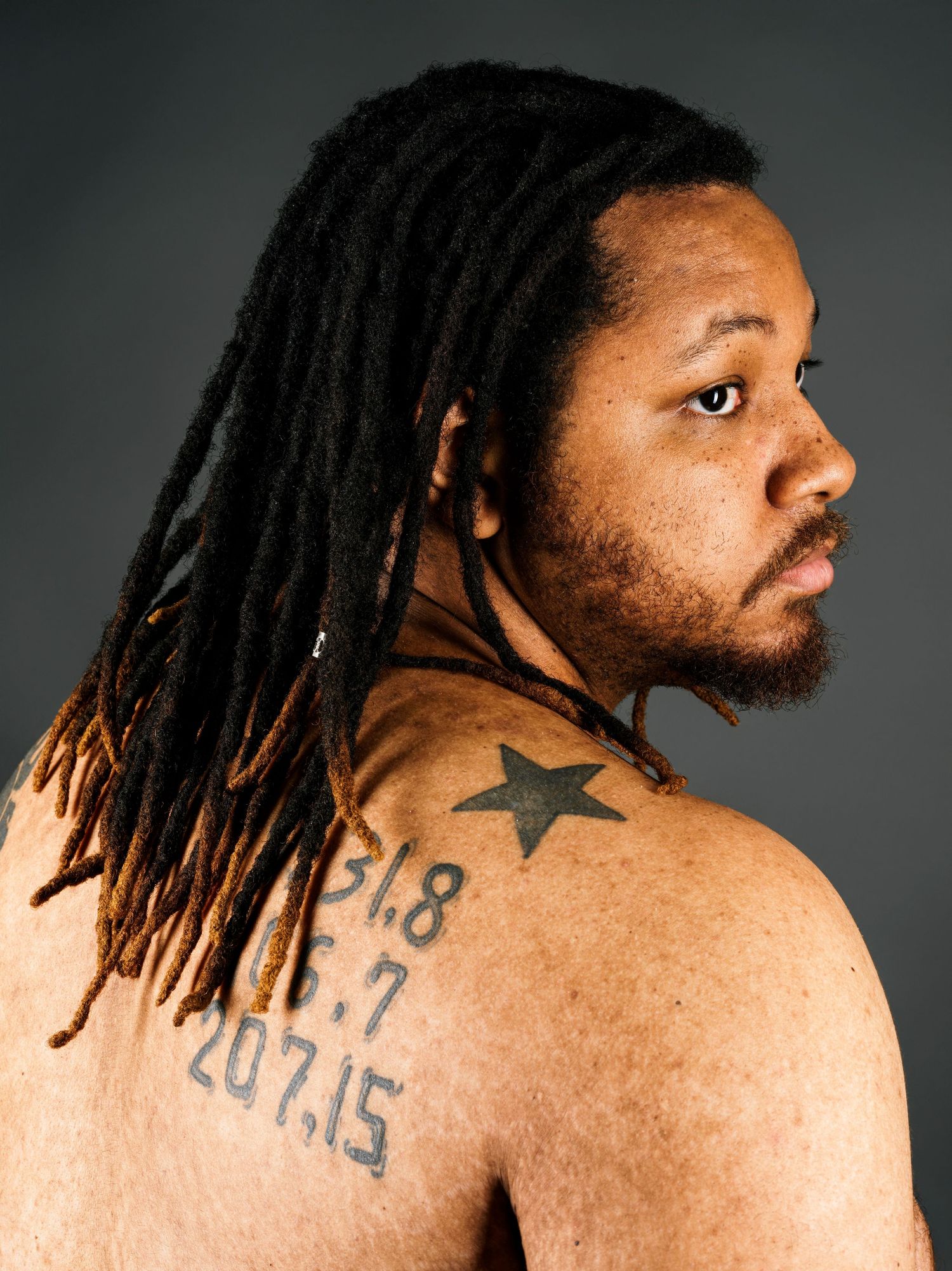 Man with dreadlocks showing back tattoos including a star and numbers on a dark background.