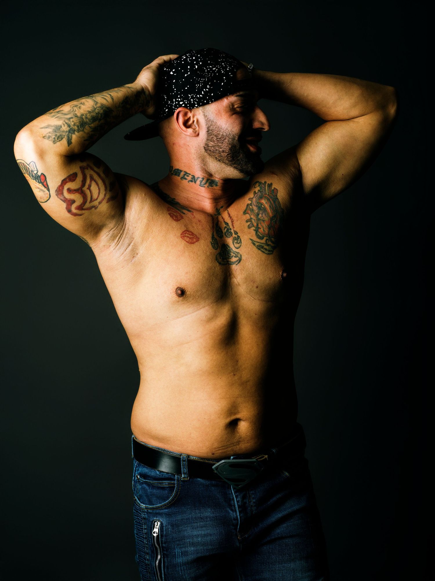 Muscular tattooed shirtless man in jeans poses with hands on head wearing a sparkly cap against dark background.