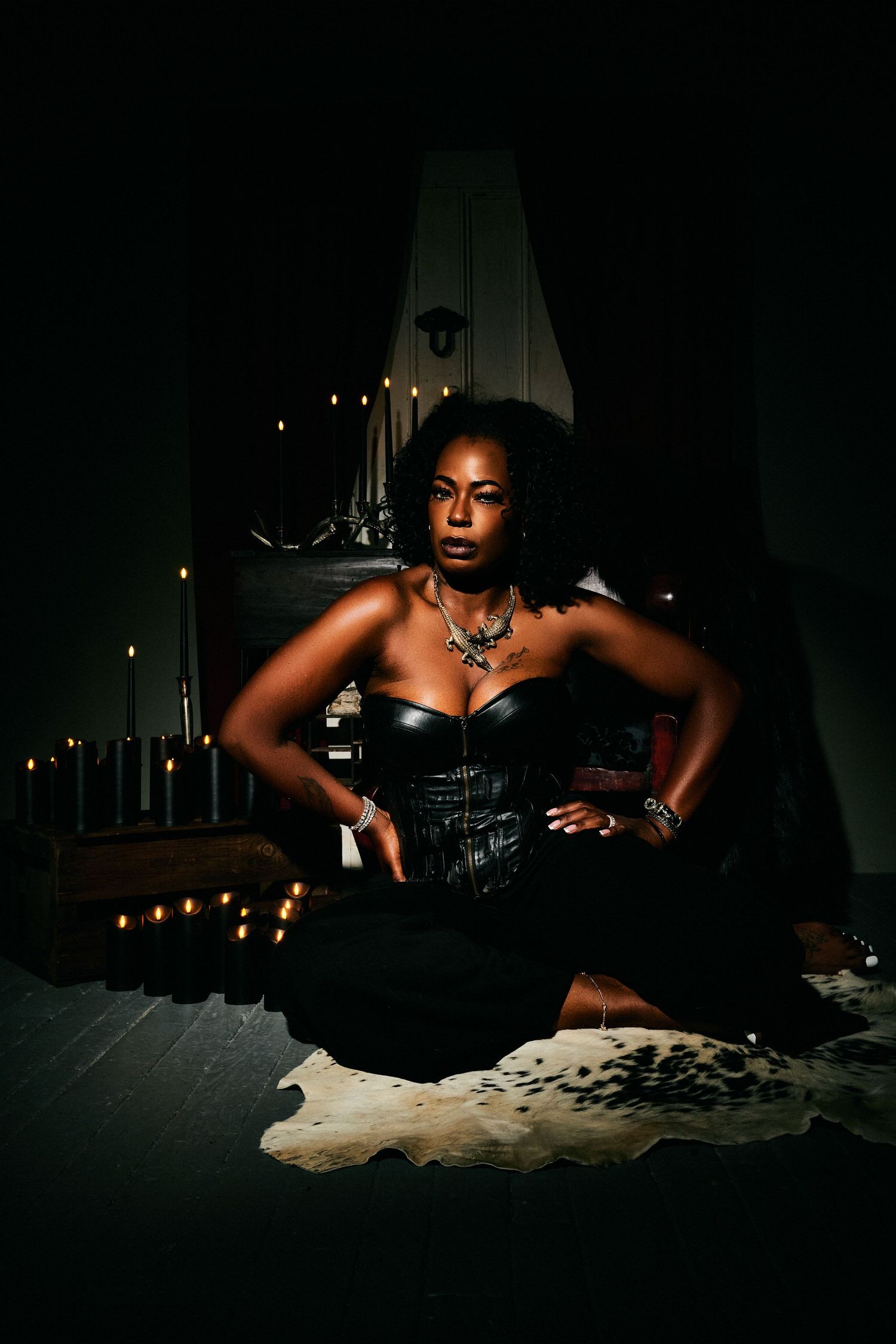 A woman in a black corset sits on a fur rug surrounded by candles in a dark, moody setting.