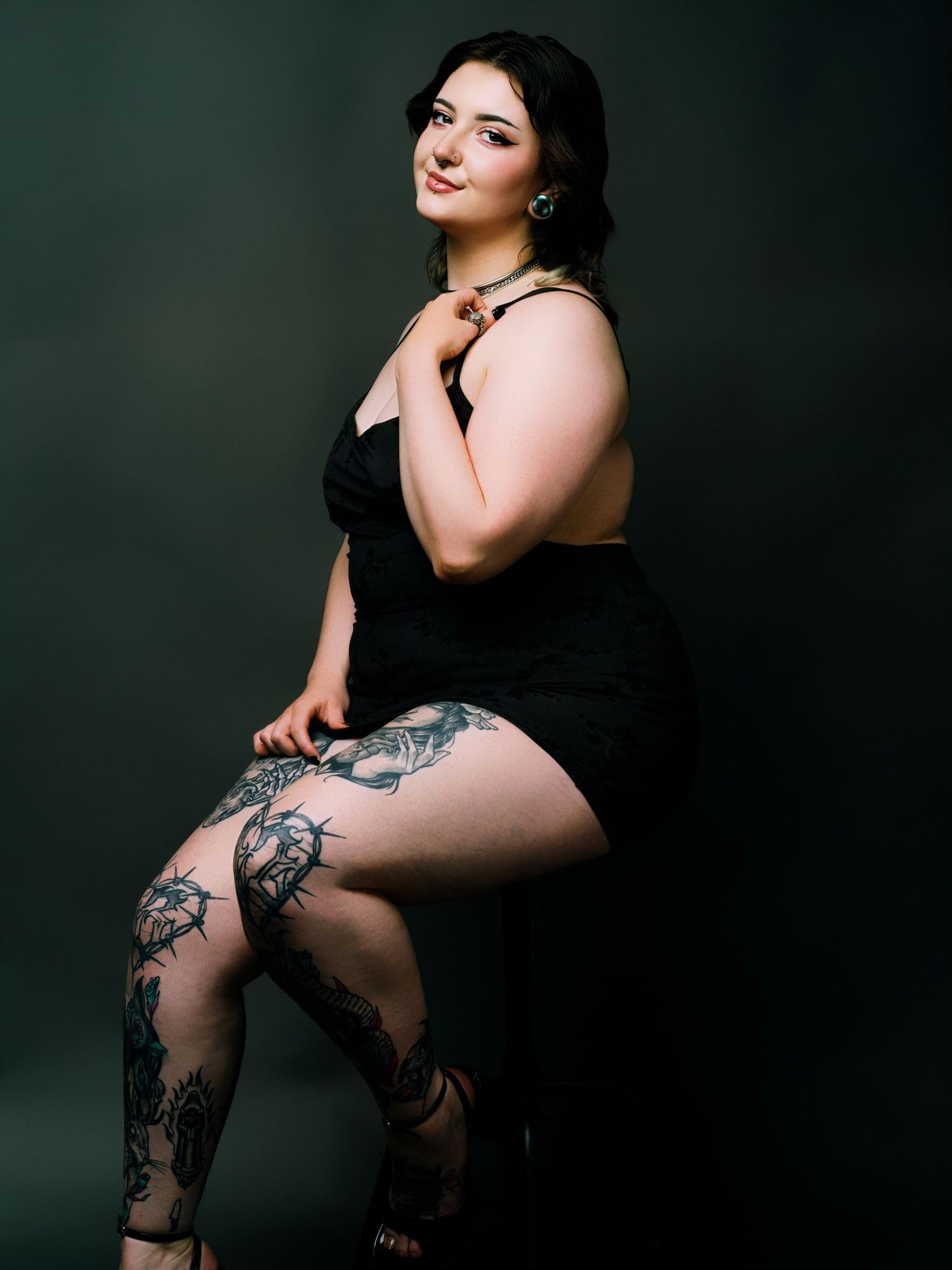 Woman in black dress with tattoos on legs sits elegantly against dark background in studio portrait.