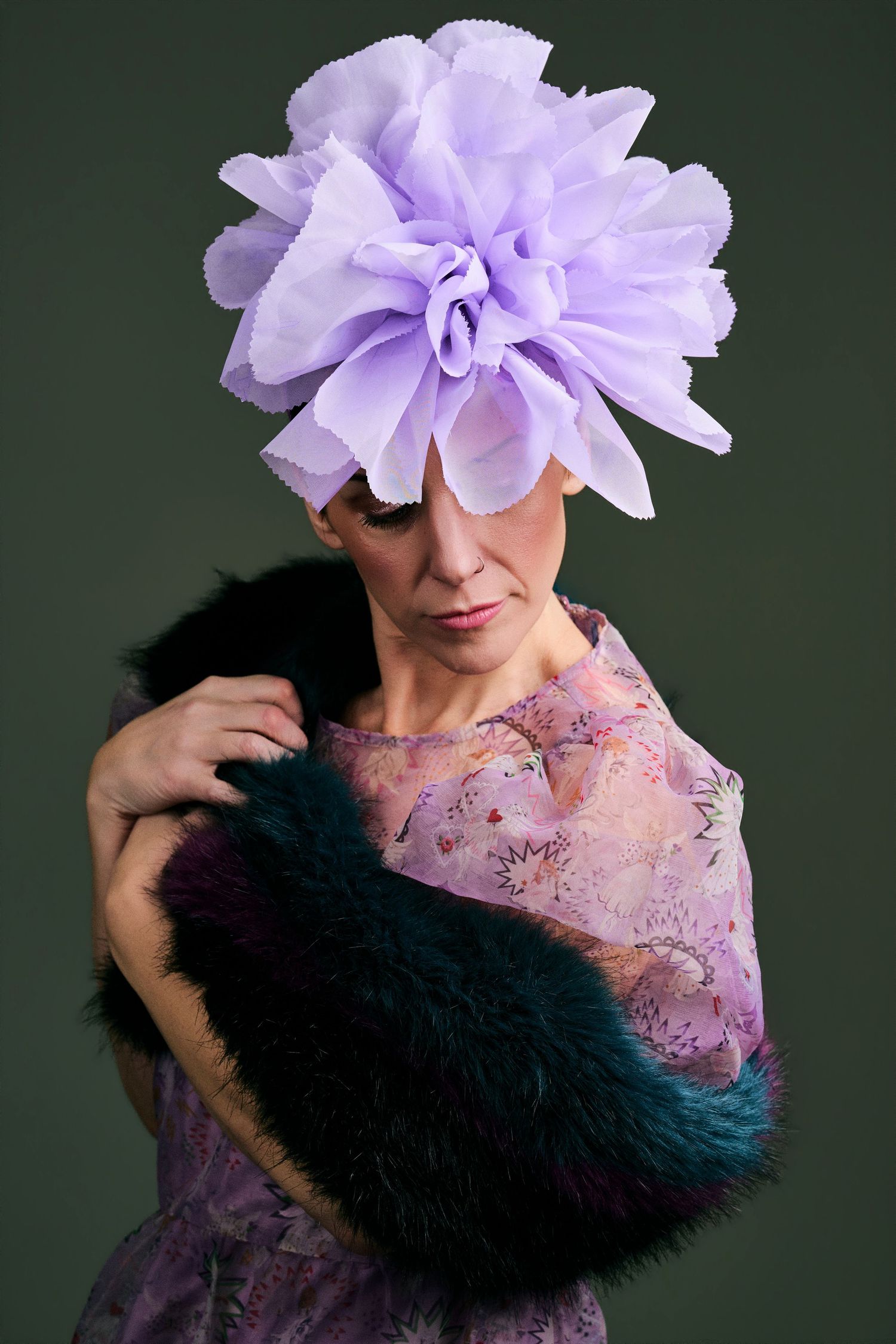 Woman in lavender floral dress wearing large purple flower fascinator and teal fur stole on dark background.