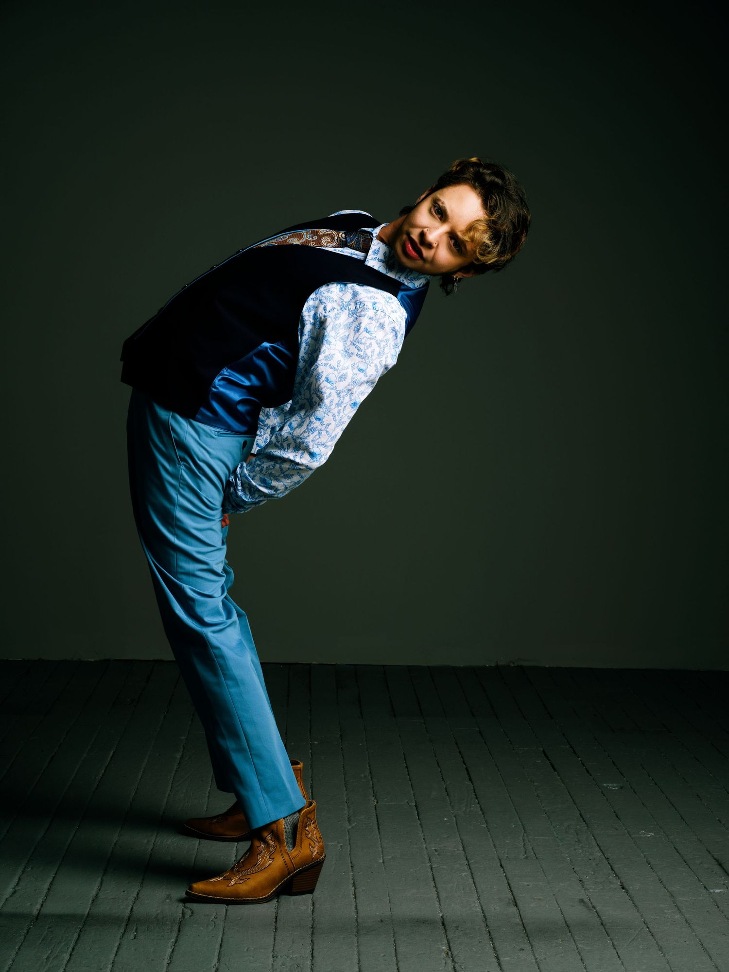 Man in blue suit vest and floral shirt poses leaning forward, wearing tan cowboy boots on wooden floor.