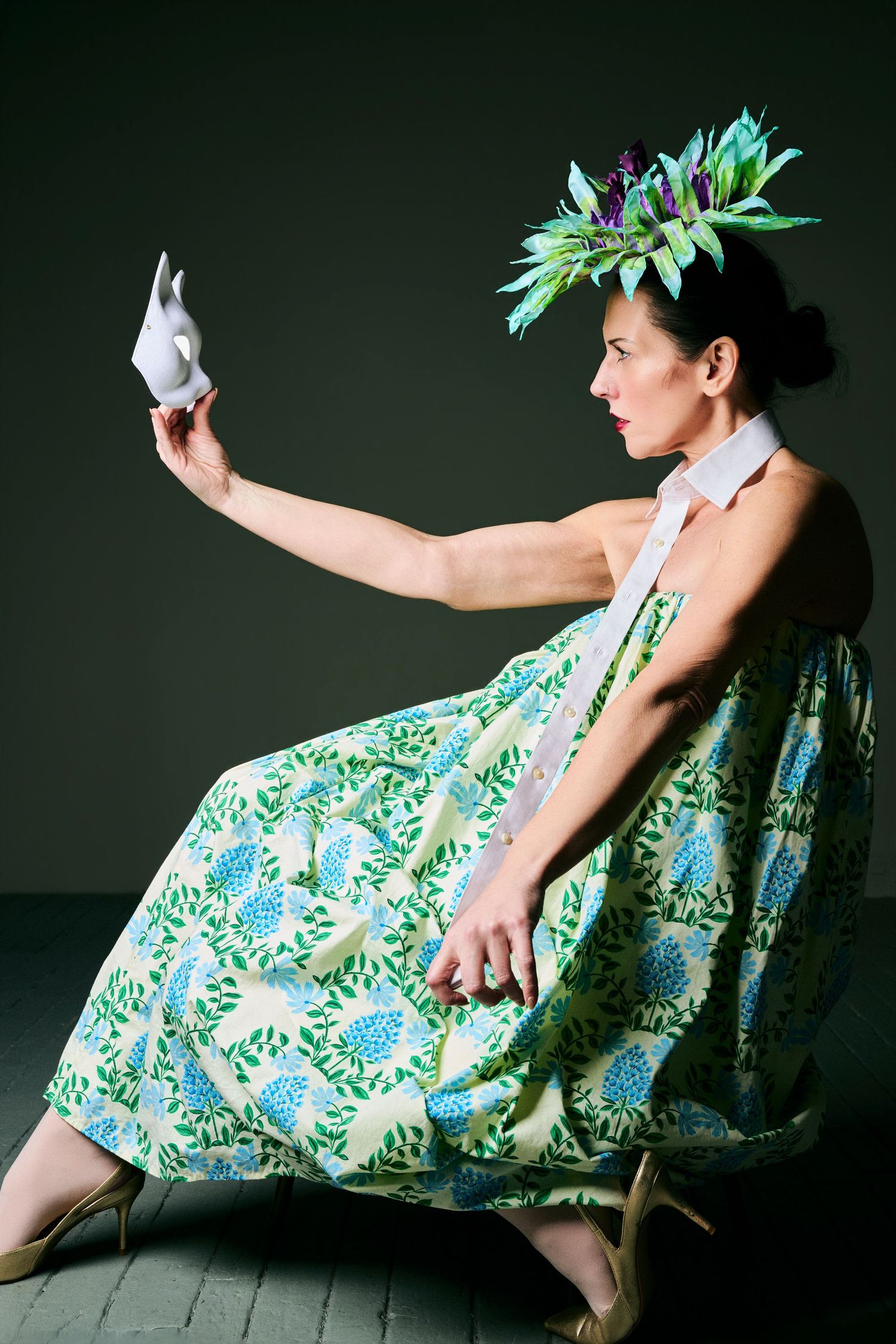 Woman in floral dress and leaf crown holds white origami bird while seated on wooden chair against dark background.