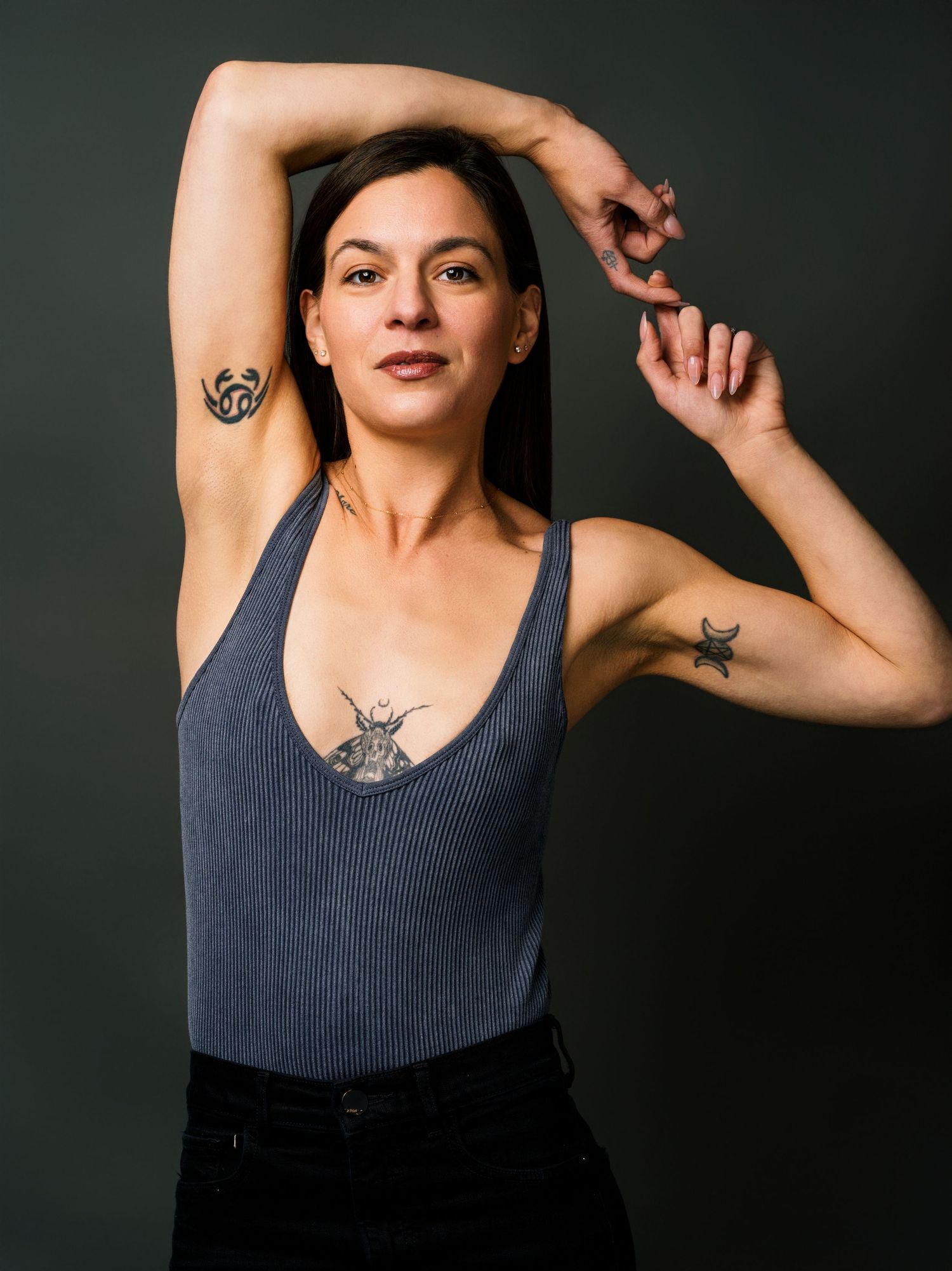 Woman with tattoos poses in gray tank top, raising arms to show armpit and bicep tattoos against dark background.