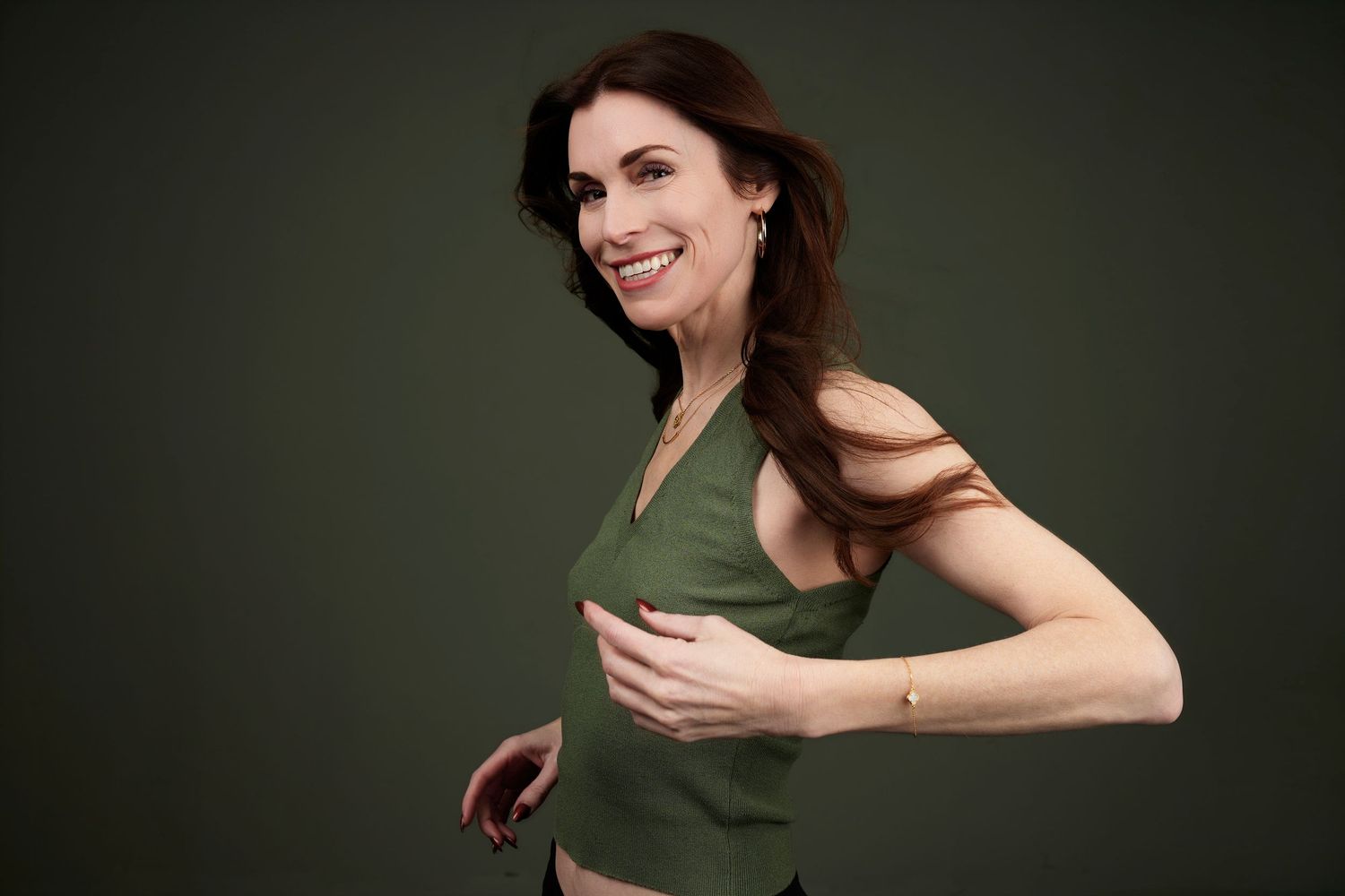 Smiling woman with brown hair wearing a green sleeveless top, posing confidently against a dark green background.