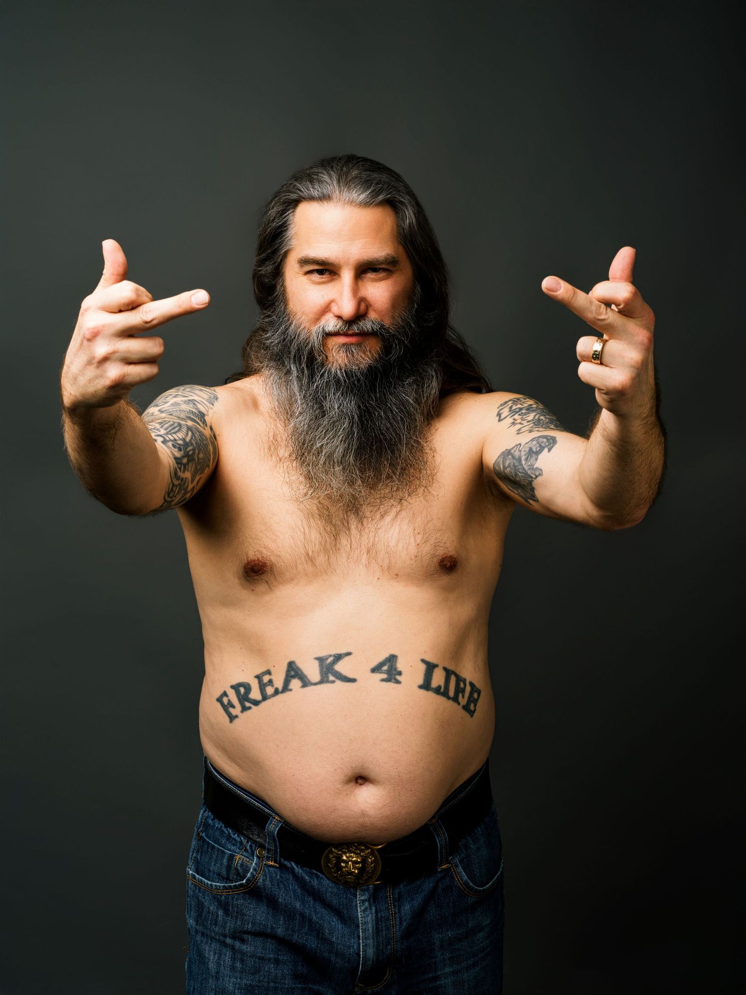 Shirtless tattooed man with long hair and beard showing rock hand gestures with FREAK 4 LIFE stomach tattoo.