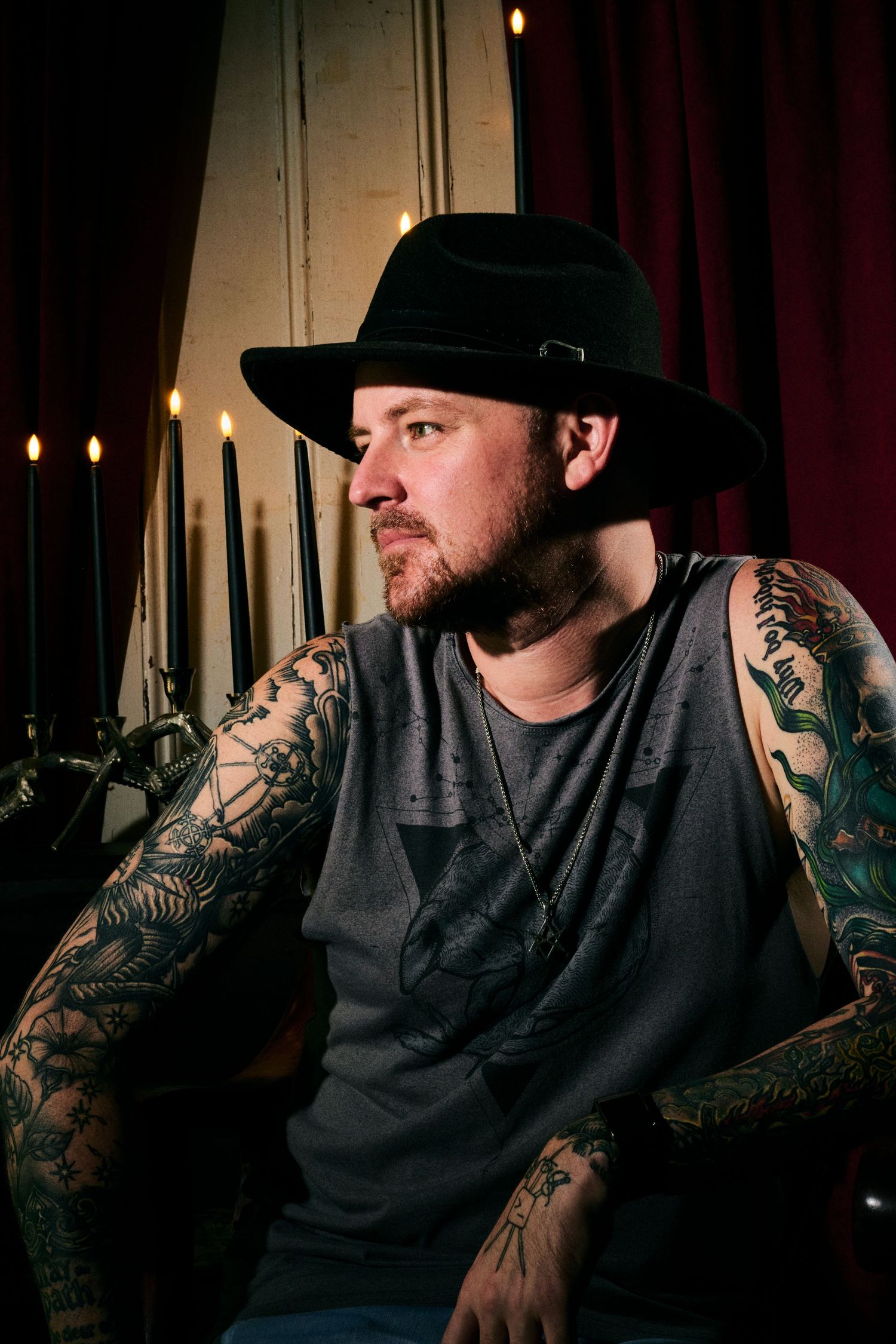 Tattooed man wearing a black fedora hat and gray t-shirt sitting in dramatic candlelit setting with red curtains.