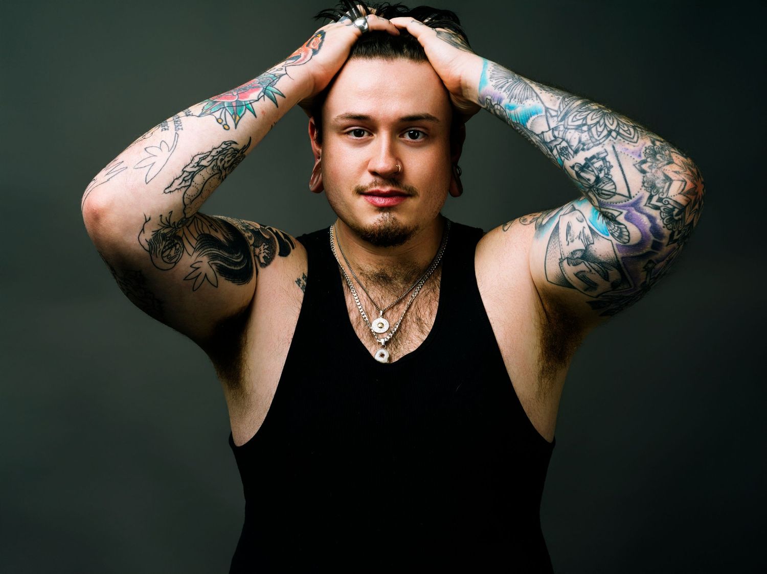 Tattooed young man in black tank top posing with hands behind head, showcasing full sleeve tattoos on both arms.