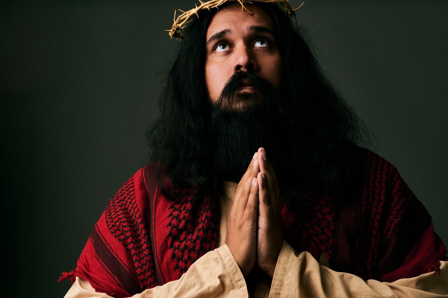 Man portraying Jesus Christ with crown of thorns, praying with hands clasped, wearing red and white robes.