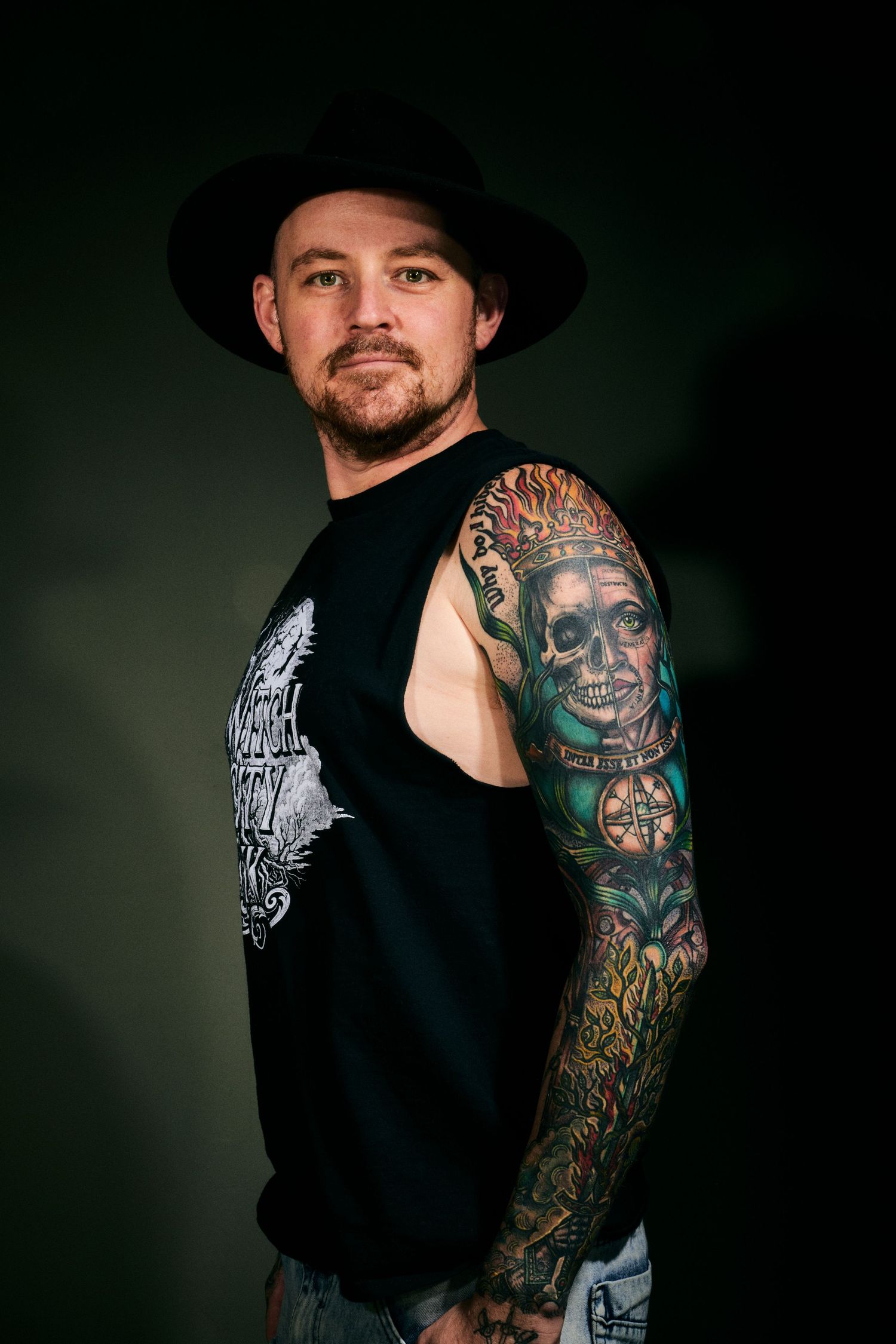 Man with black hat and colorful skull tattoo sleeve posing in black sleeveless shirt against dark background.