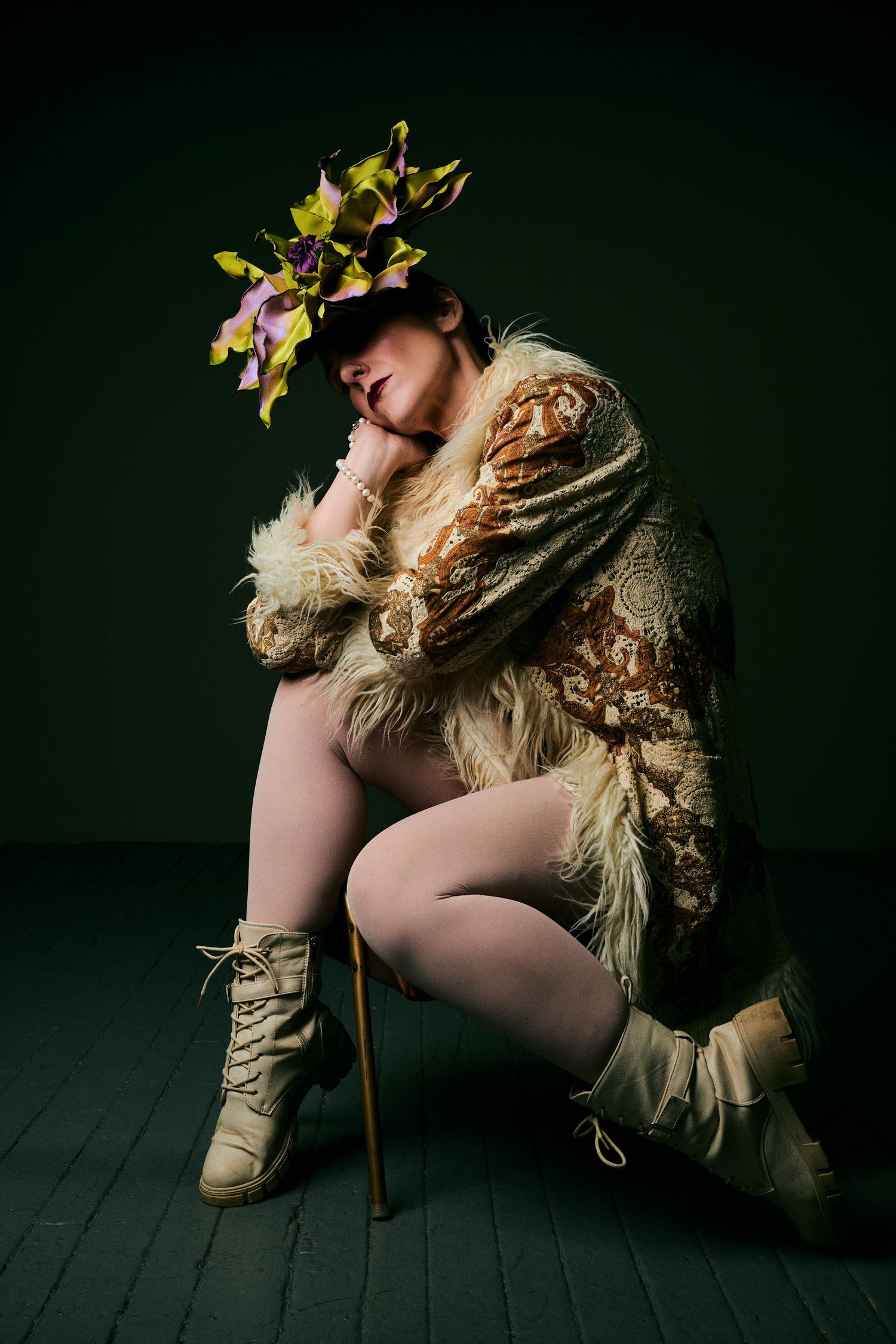 Person in fur coat and floral headpiece posing dramatically on dark green background with cane and white boots.