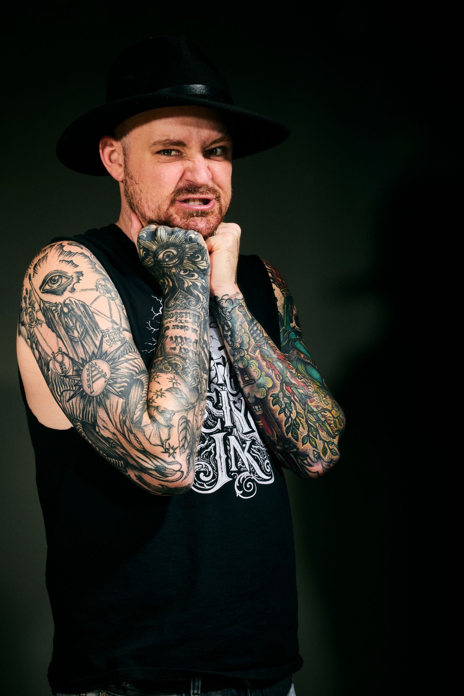 Tattooed man in black hat and sleeveless shirt posing with fist raised, showcasing full sleeve tattoos on dark background.