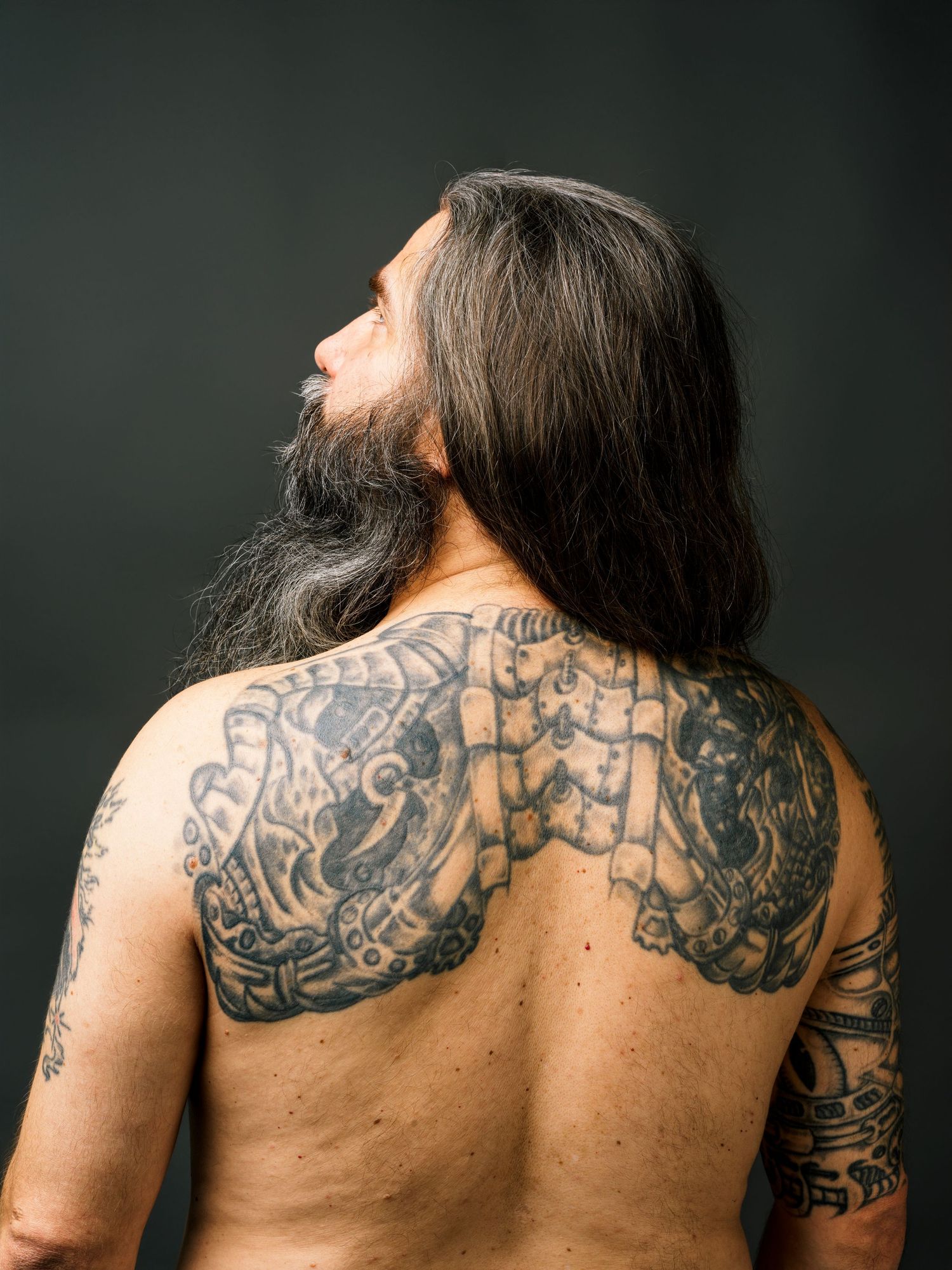 Tattooed man with long hair and beard showing detailed back tattoo artwork on dark background.