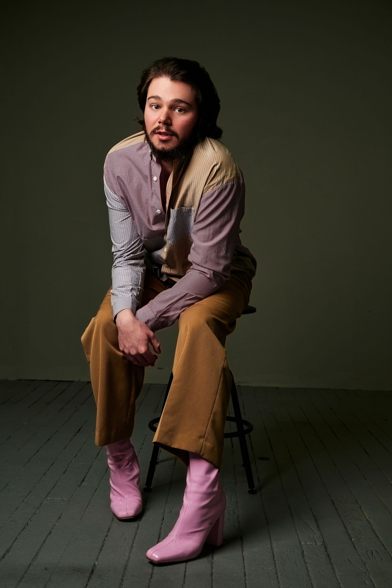 Young man sitting on stool wearing pink cowboy boots, tan flared pants, and striped shirt in studio setting.