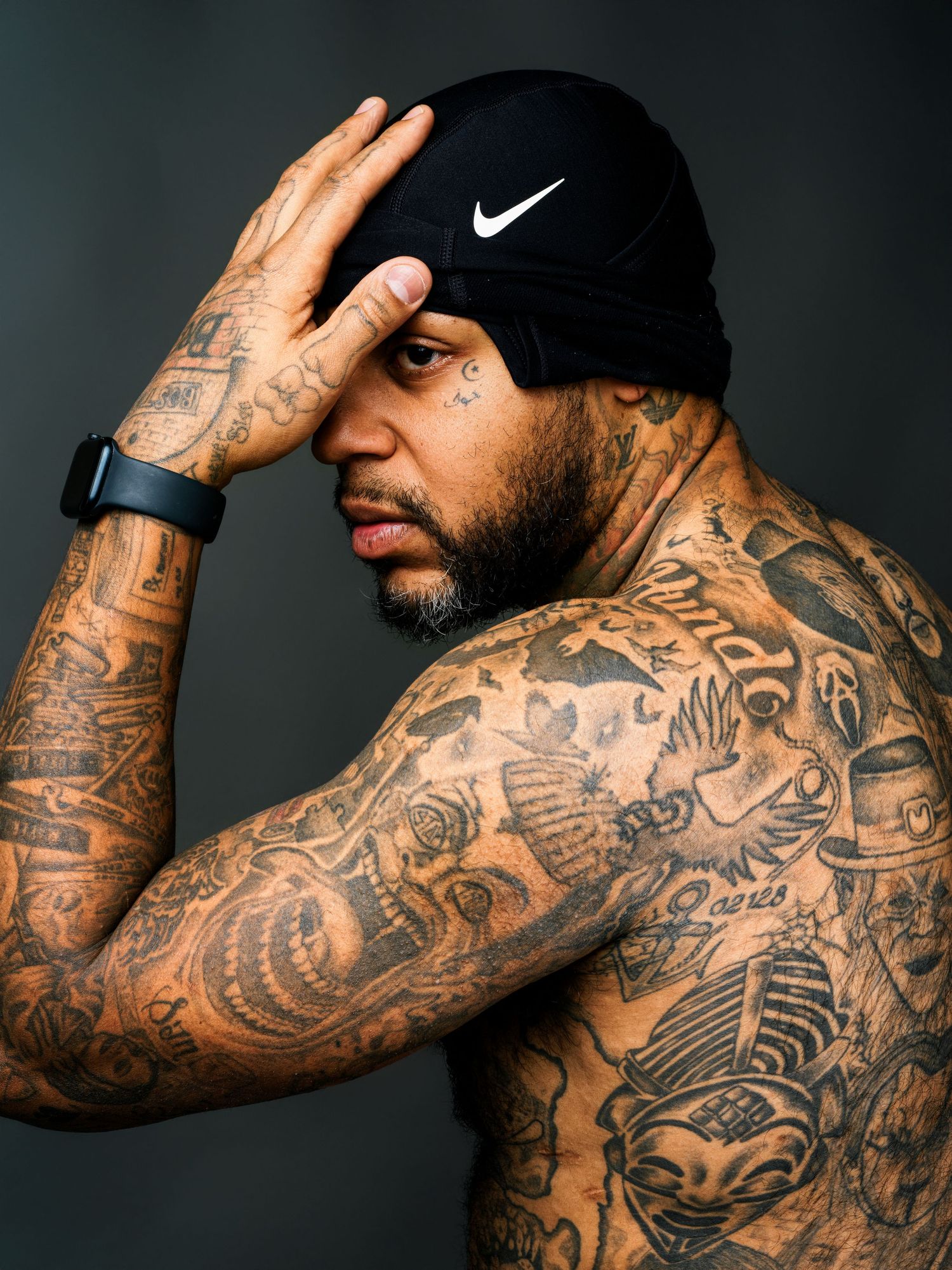 Heavily tattooed shirtless man wearing a black Nike beanie and smartwatch poses against a dark studio background.