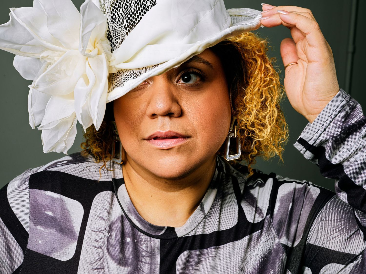 Woman in white floral hat with mesh brim, curly hair, wearing patterned gray top, posing confidently.
