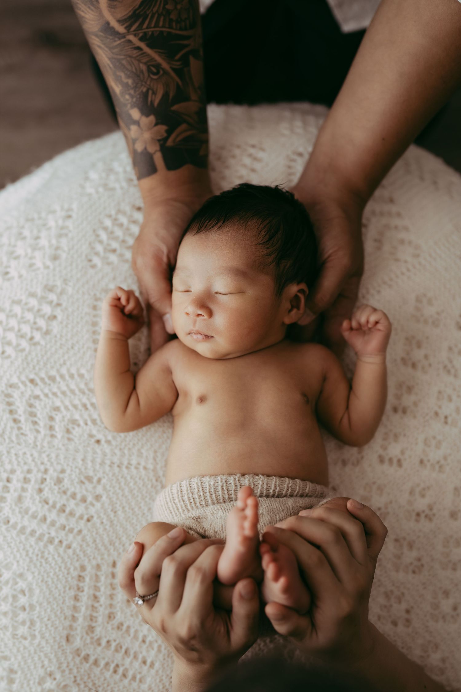 A sleeping newborn baby lies peacefully while hands with tattoos gently support them.