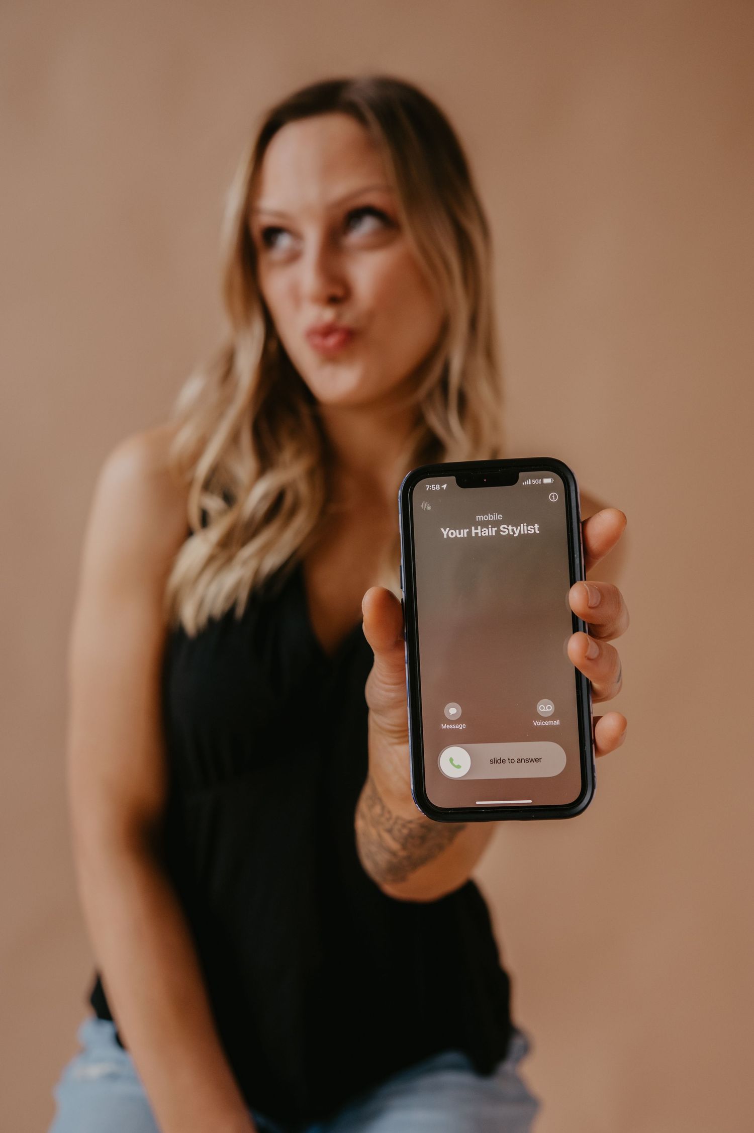 Woman with wavy blonde hair holding up an iPhone showing an incoming call from Your Hair Stylist while making a playful pouty face.