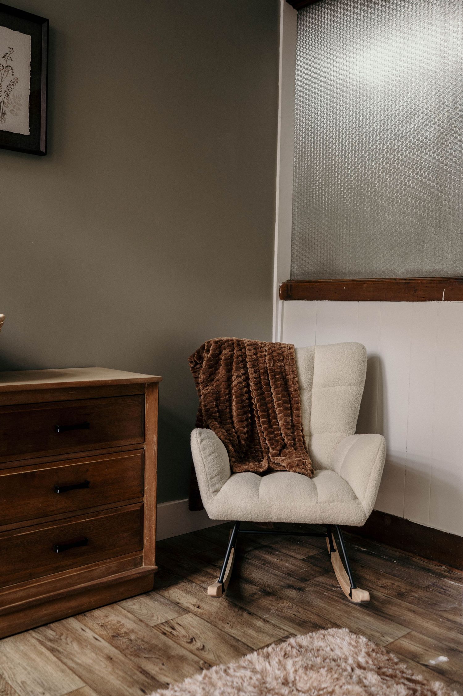 Cream tufted rocking chair with a brown faux fur throw beside a wooden dresser in a cozy room with olive walls and wood flooring.