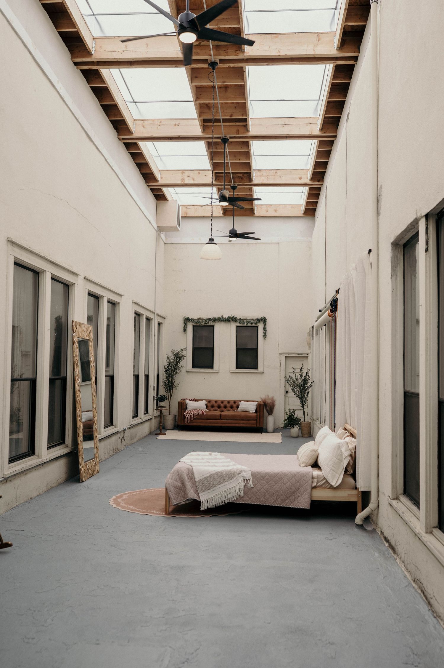 Bright loft studio with skylights, ceiling fans, a rustic bed, leather sofa, tall windows and a gold floor mirror on a concrete floor.
