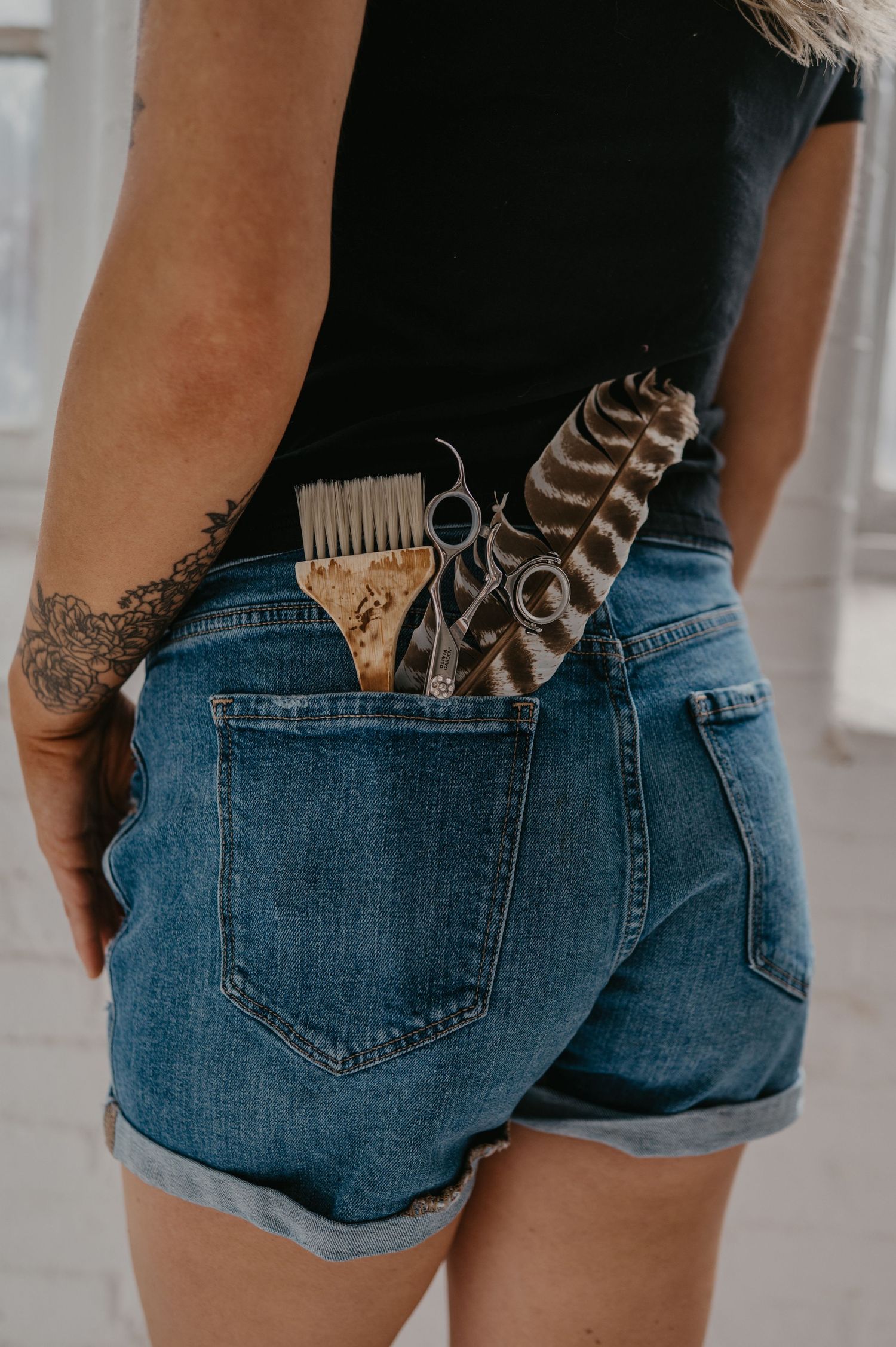 Hairstylist in black tee and denim shorts with a tattooed arm, carrying a color brush, shears and a feather in her back pocket.