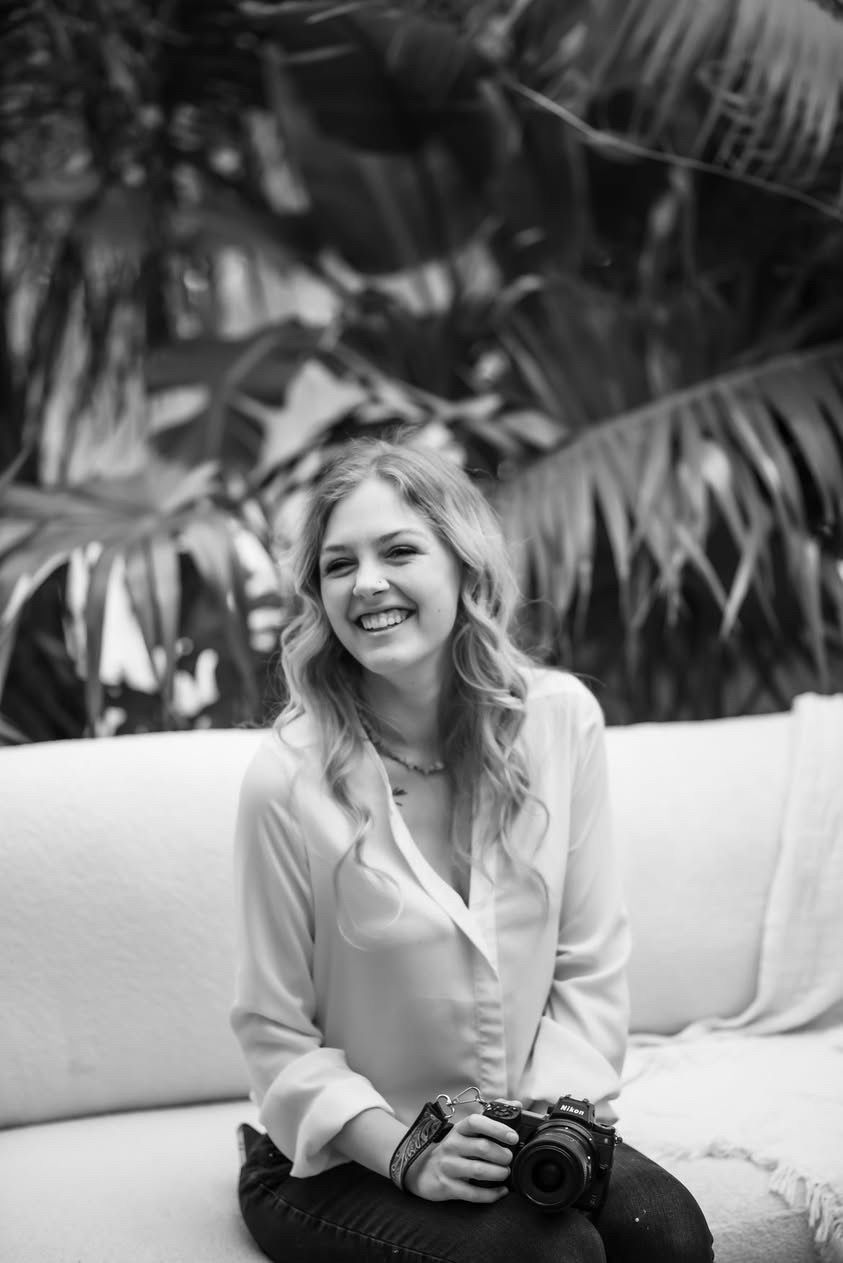 Black and white portrait of someone smiling while sitting with a camera on a white couch with palm leaves in background.