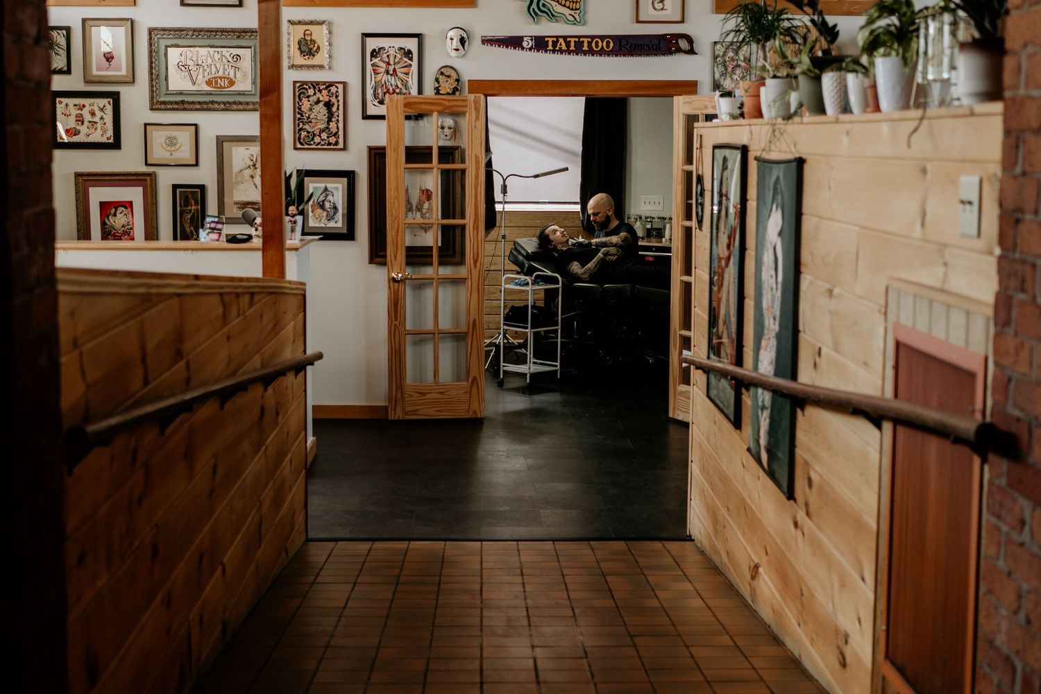 Tattoo shop entrance with wood-paneled walls, framed art, a glass-paned door and an artist seated in the studio beyond a tiled hallway.