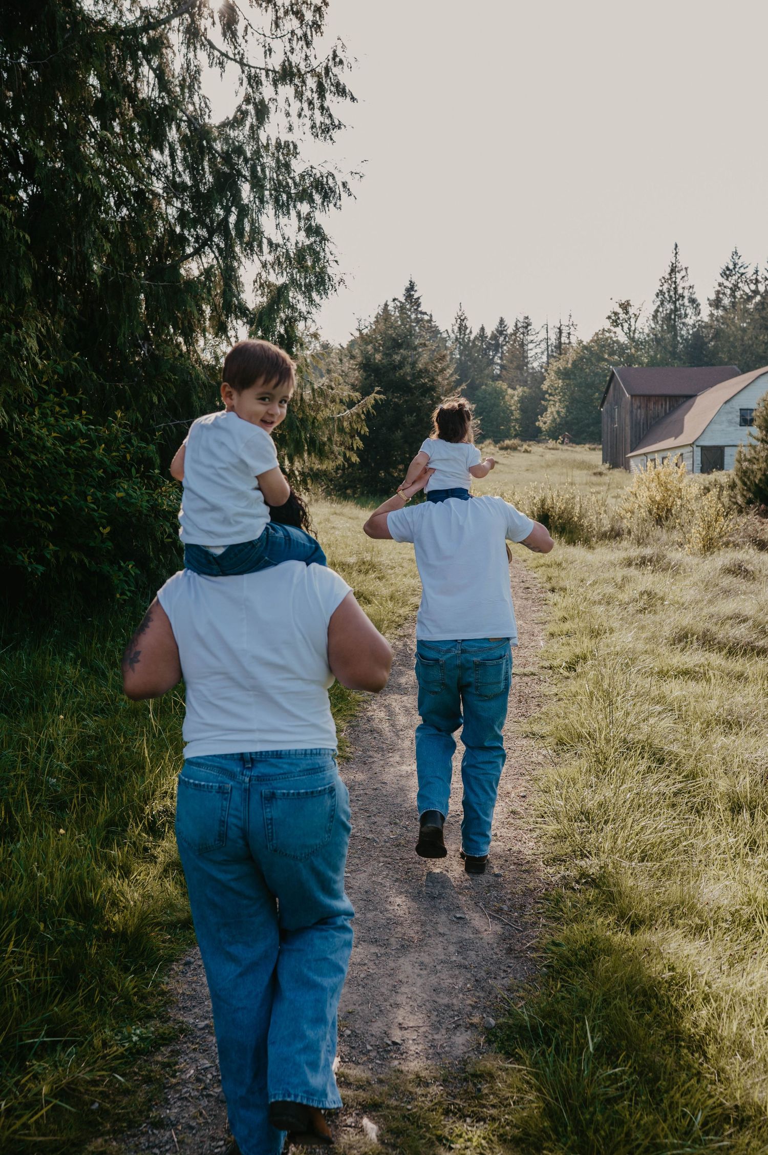 Two adults in white shirts and jeans walk down a grassy dirt path carrying children on their shoulders toward a rustic barn.