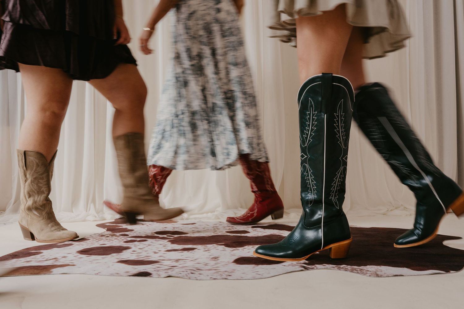Three women in skirts and dresses wear tan, red, and black western cowboy boots while standing on a cowhide rug.