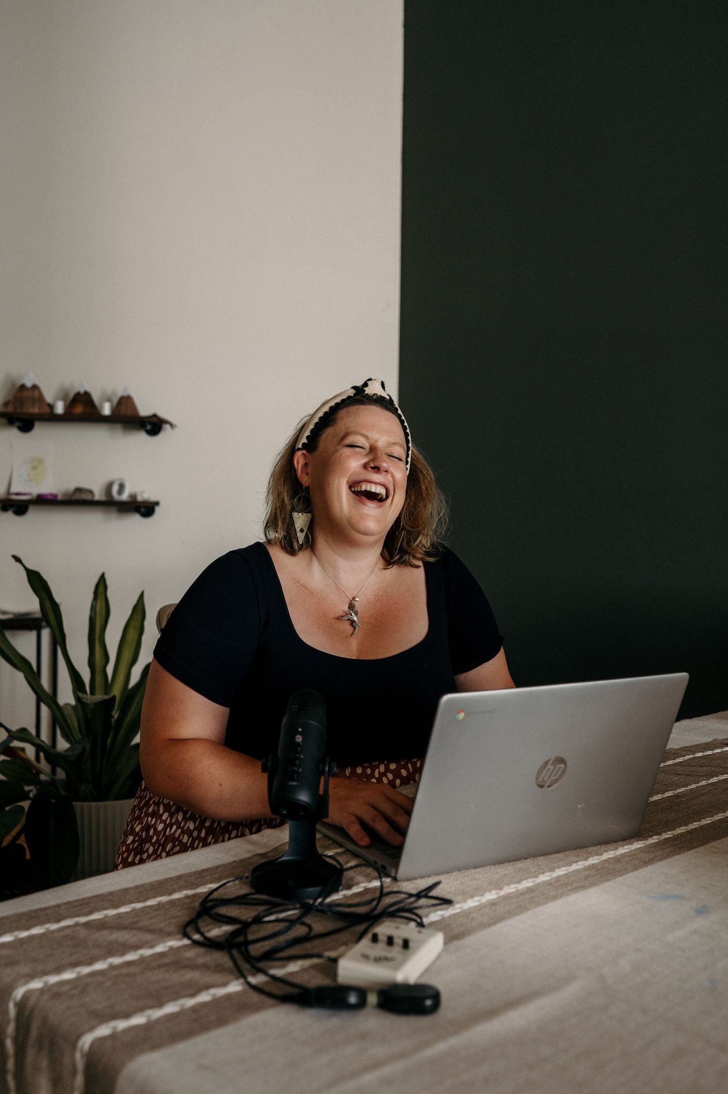 Smiling woman laughing at a table with a laptop and podcast microphone, wearing a black top and patterned headband in a bright room.