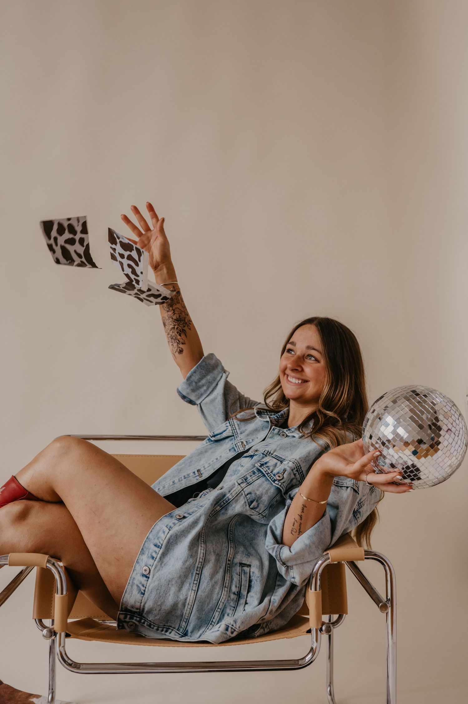 Smiling woman in an oversized denim jacket sits in a modern chair, tossing patterned cards while holding a mirrored disco ball.
