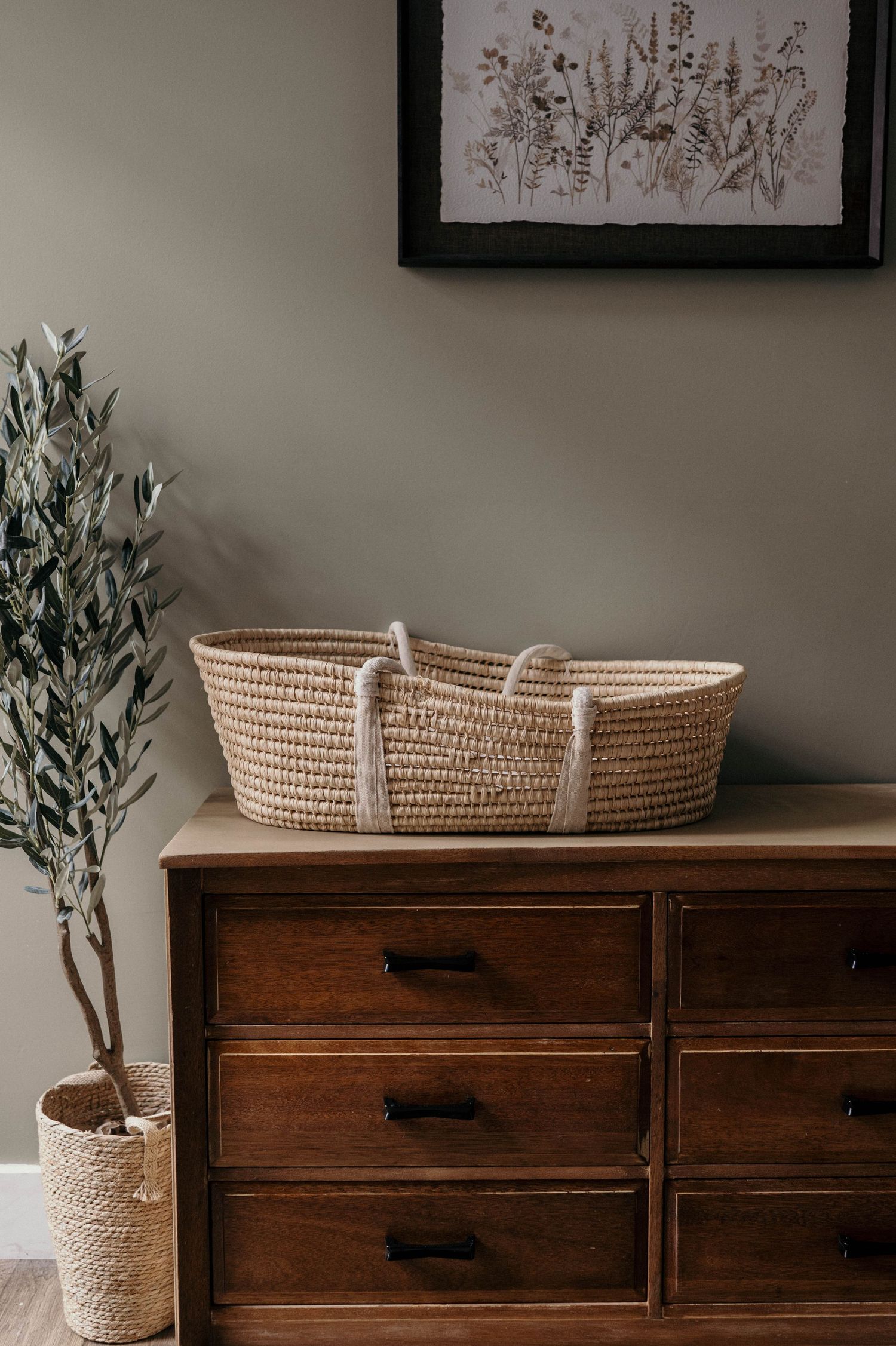 Woven rope Moses basket with cream handles on a vintage wooden dresser beside a potted olive tree and a framed botanical print.
