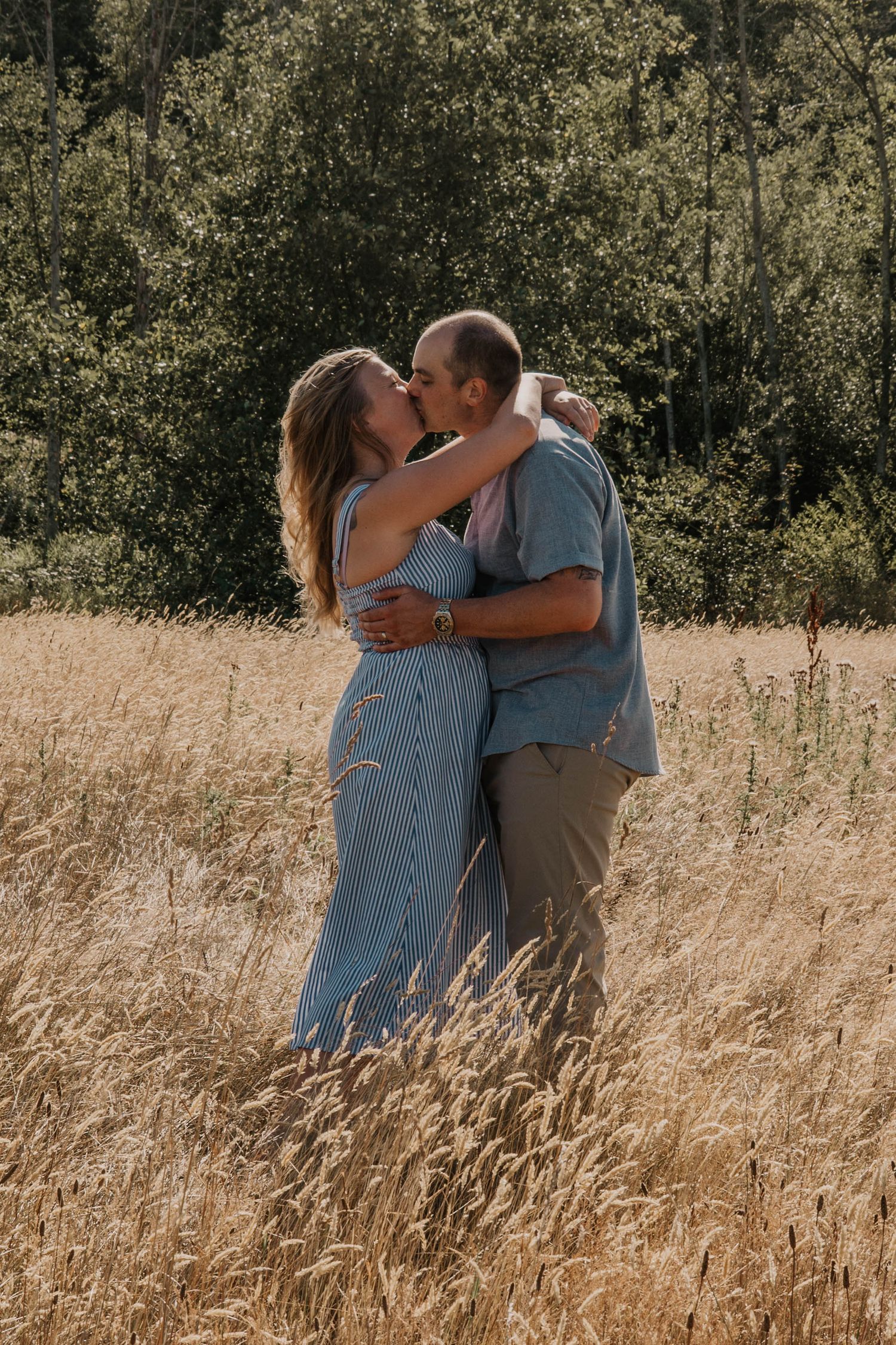 Couple in casual summer clothes embracing and kissing in a tall golden grass field with green trees in the background.