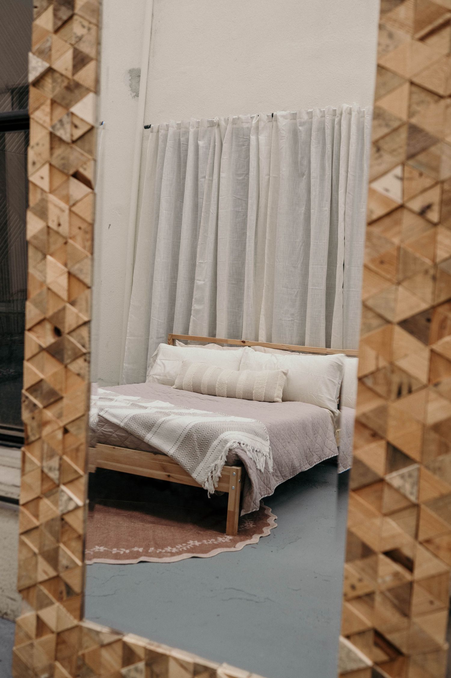 Geometric wooden mosaic framed mirror reflecting a rustic wood-frame bed with white linens, a knit throw, and a round woven rug.