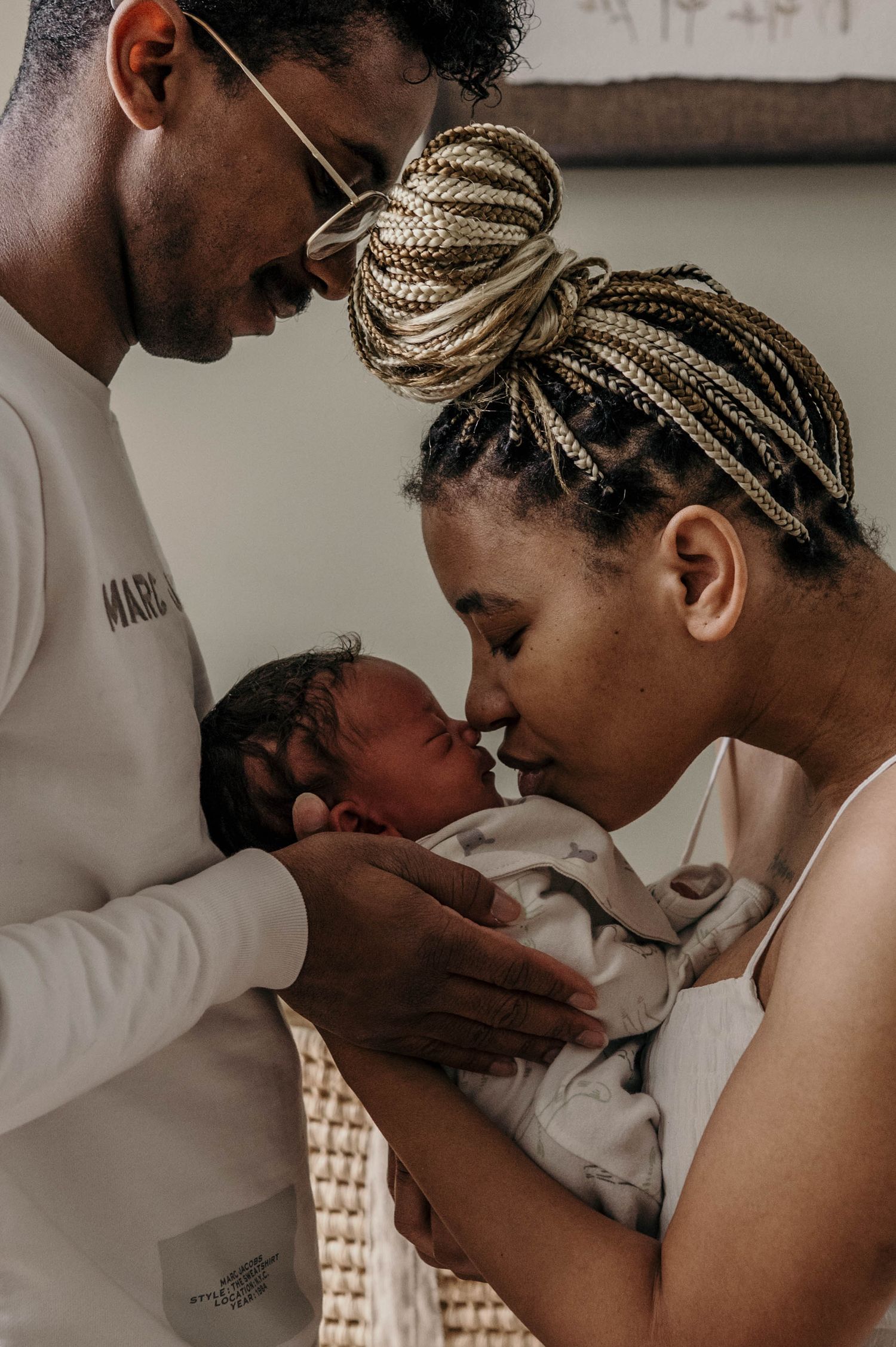 Two parents tenderly embrace and gently kiss their sleeping newborn baby wrapped in a soft blanket during an intimate family moment at home.