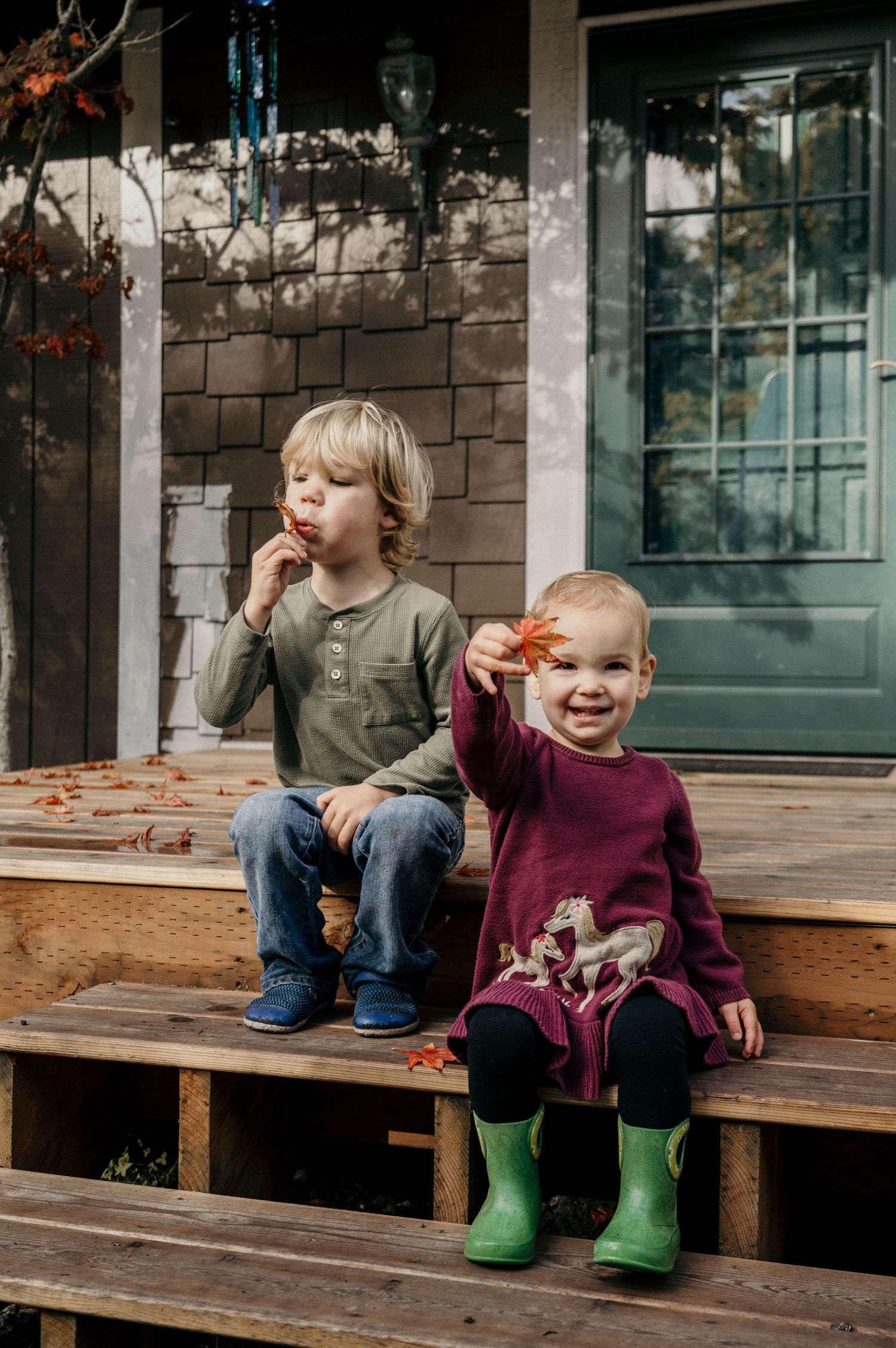 Two young children in green rain boots and cozy fall clothes sit on wooden porch steps holding colorful autumn leaves.