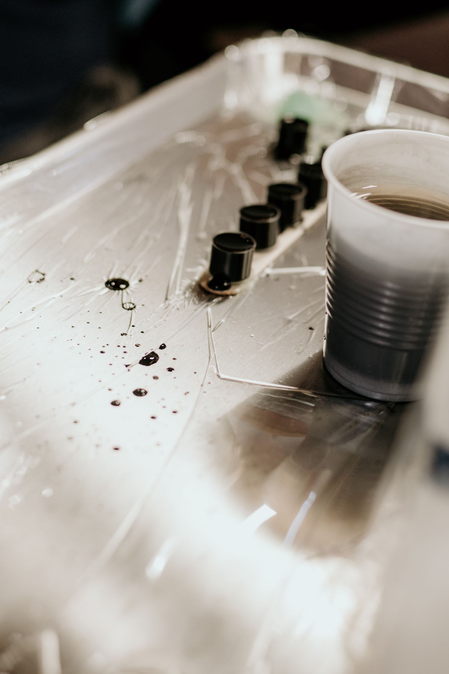 Small black ink caps and a plastic cup of dark ink sit on a glossy, splattered tray covered in clear protective film.
