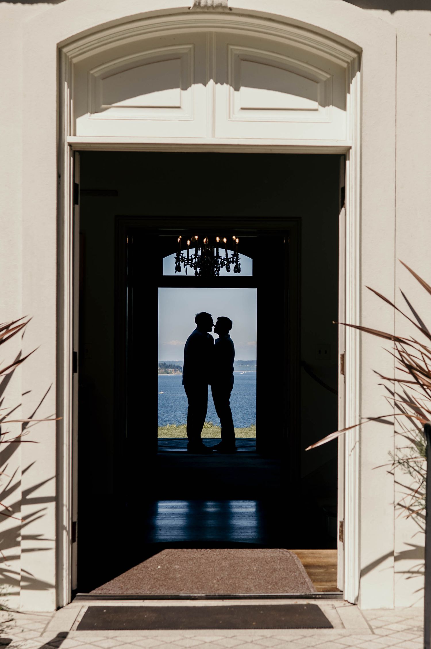 Silhouetted couple embracing in a grand arched doorway, framed by a chandelier and a sunlit waterfront view beyond the dark interior.