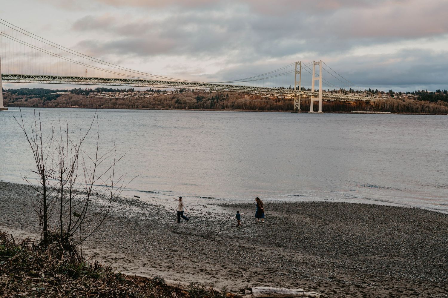 Three people walk along a pebbly shoreline beside calm water with a long suspension bridge spanning the waterway under a cloudy sky.