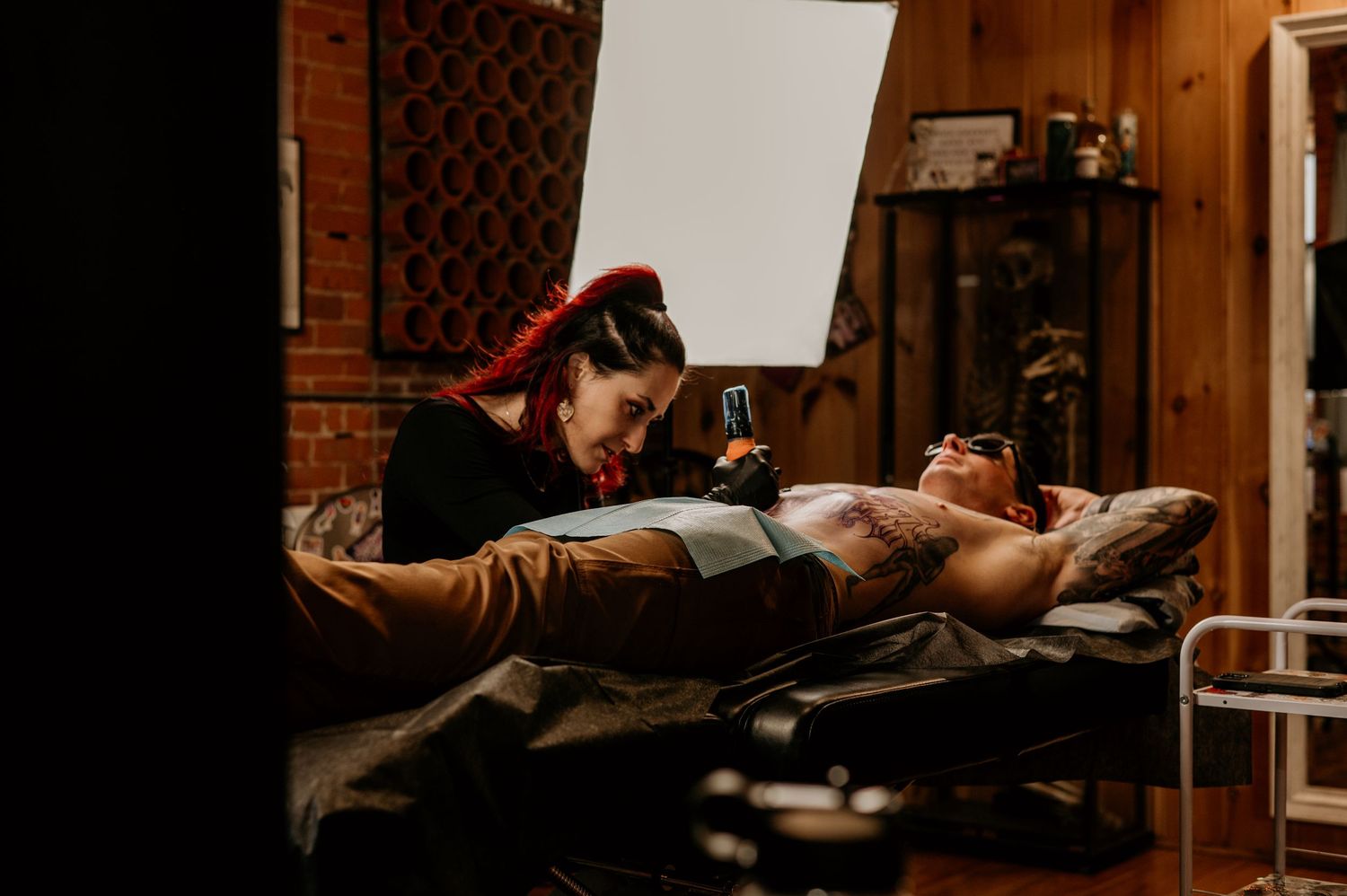 Tattoo artist with red hair focuses on inking a client lying on a black tattoo bed in a rustic studio with brick walls and wood paneling.