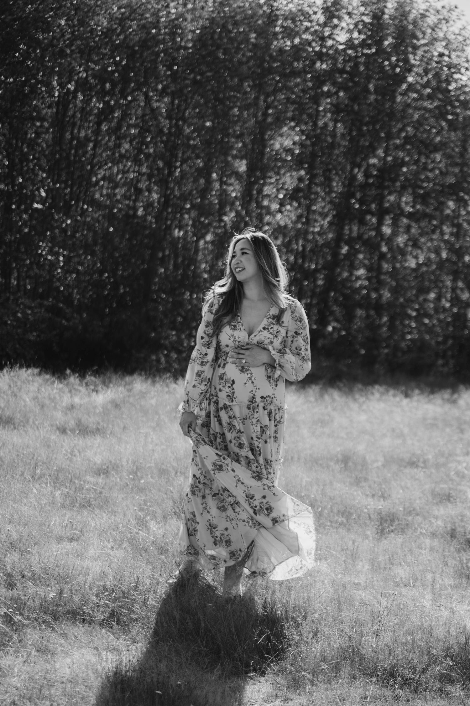 Black and white photo of a smiling woman in a flowing floral maxi dress walking through a sunlit grassy field with trees behind her.