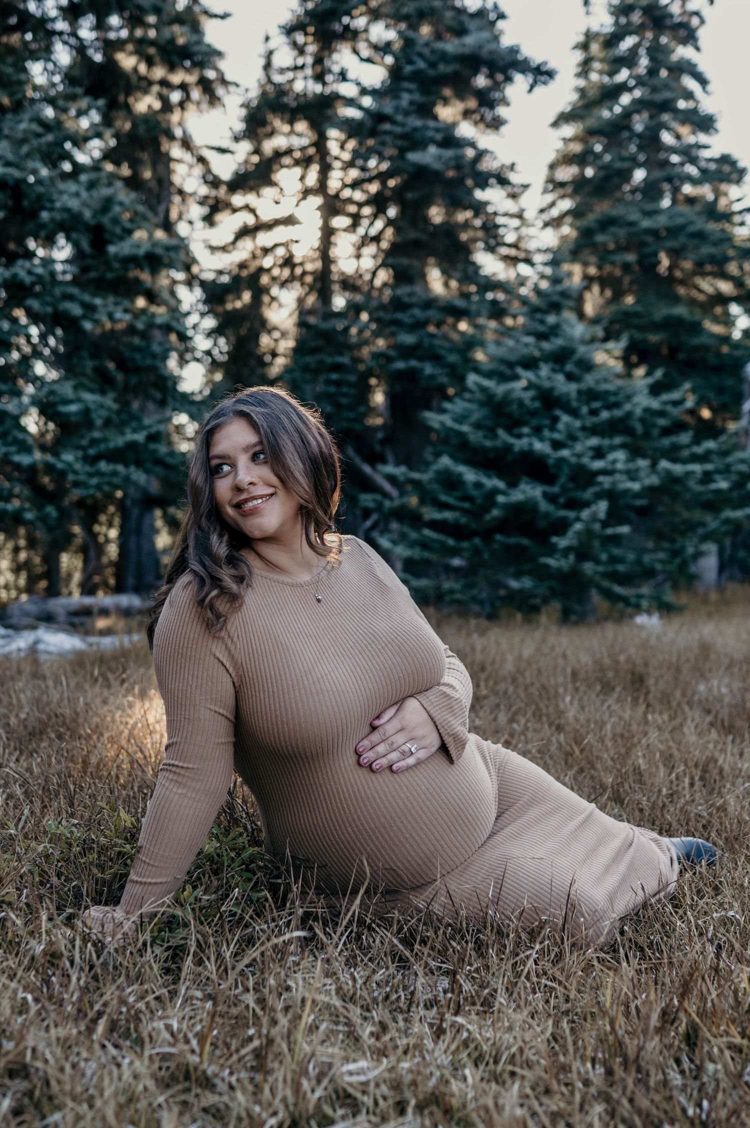 Pregnant woman in a tan ribbed dress sits smiling on dry grass, hand on her belly, with evergreen trees behind her in a maternity photo.